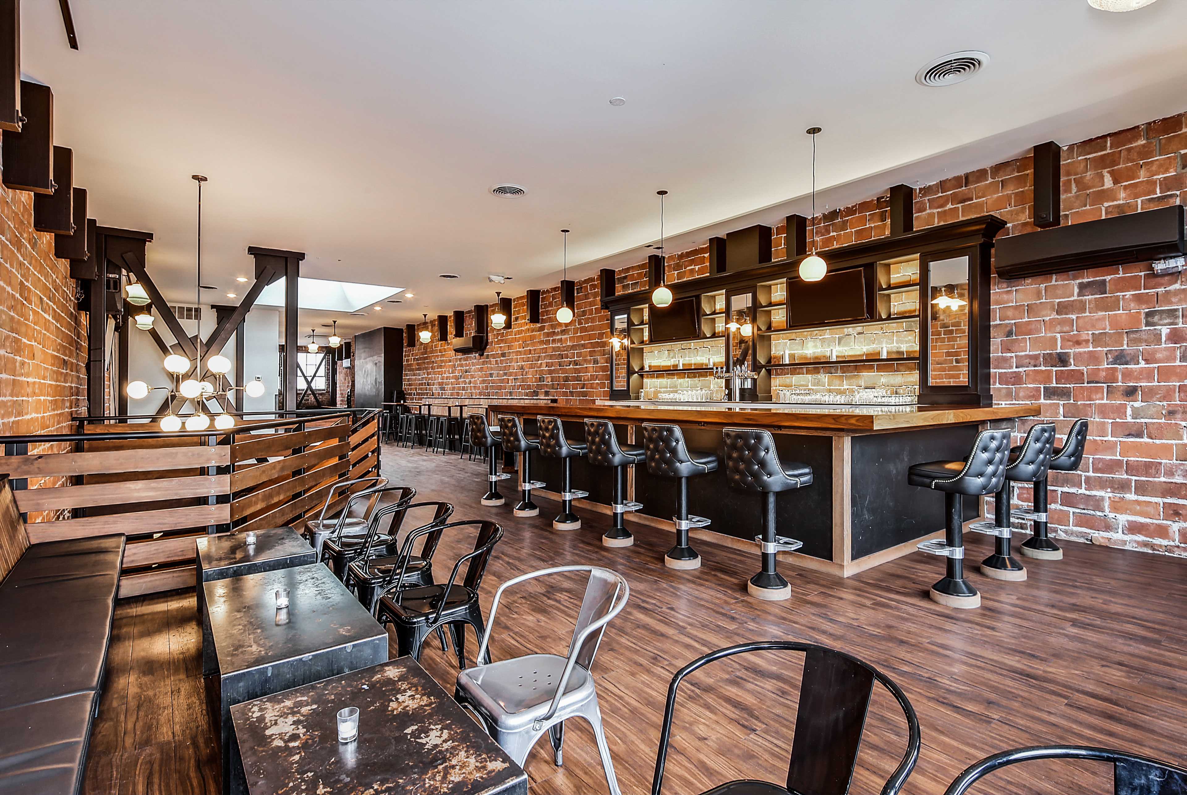 A modern bar with a brick wall, featuring black stools, a countertop, and tables in a spacious interior.