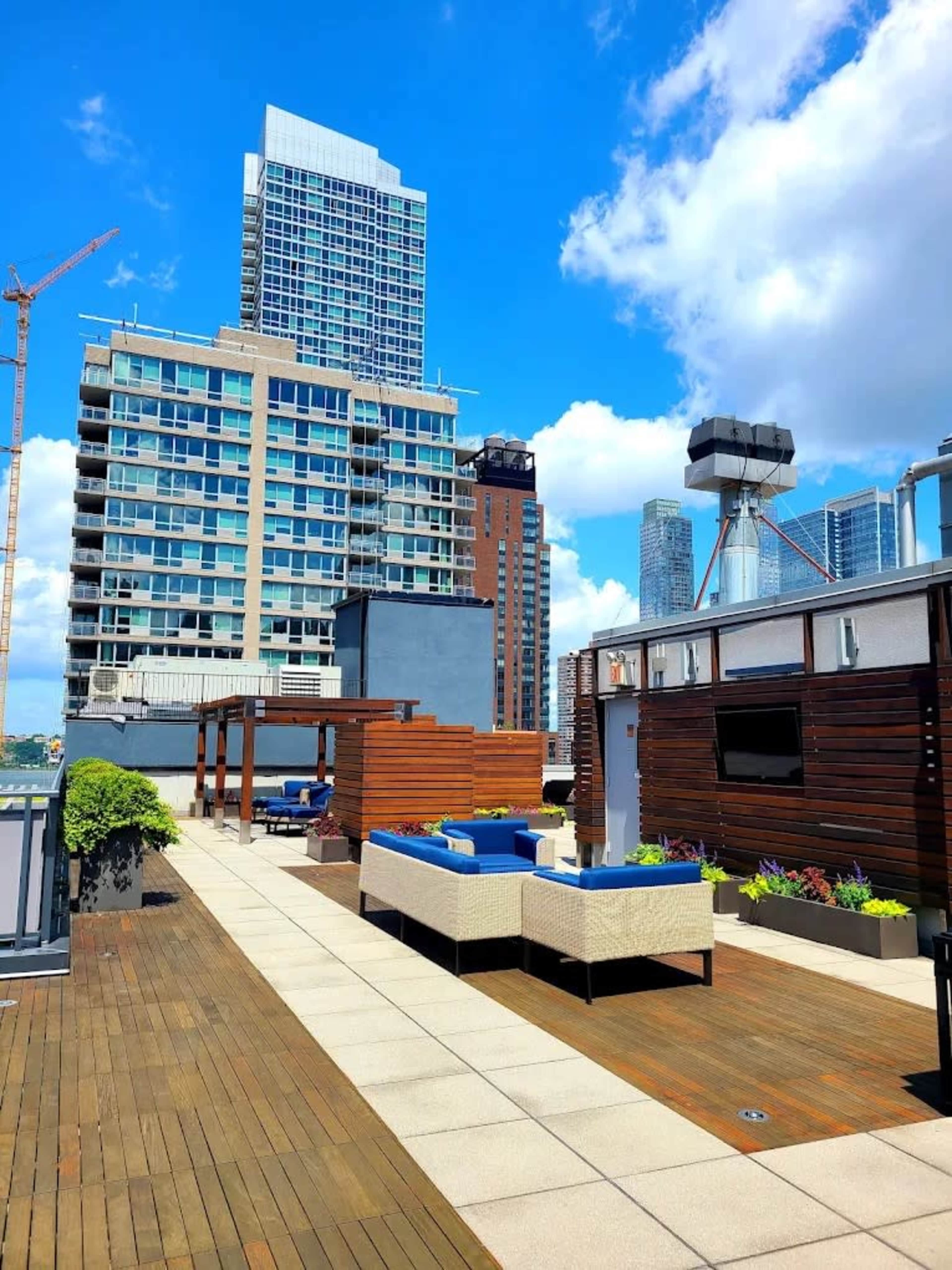 The image shows a rooftop terrace with wooden decking, lounge seating, and city skyscrapers in the background under a blue sky with scattered clouds.