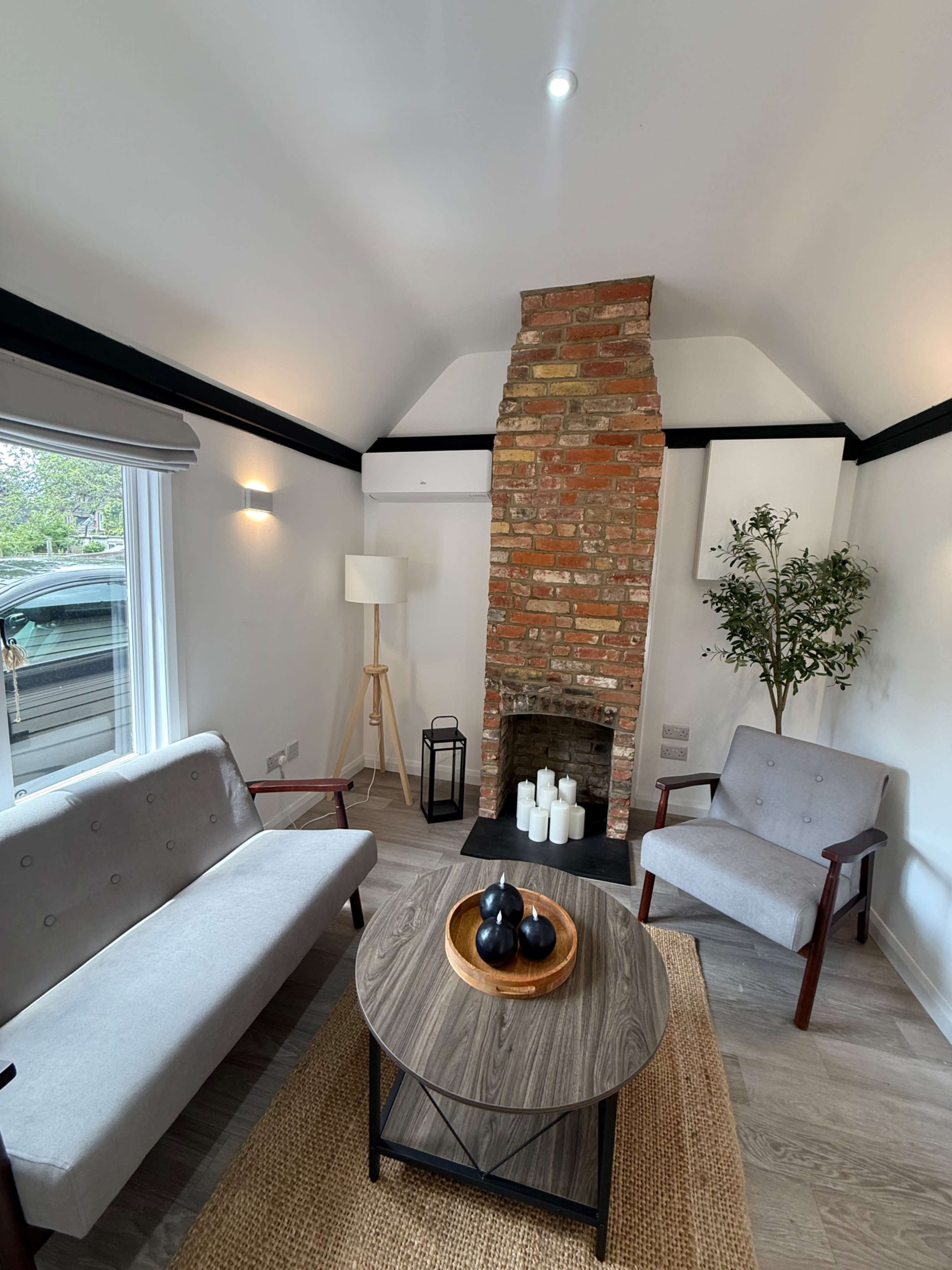 The image shows a modern living room featuring a brick fireplace, two grey armchairs, a round wooden coffee table with decorative items, and natural light coming through a window.