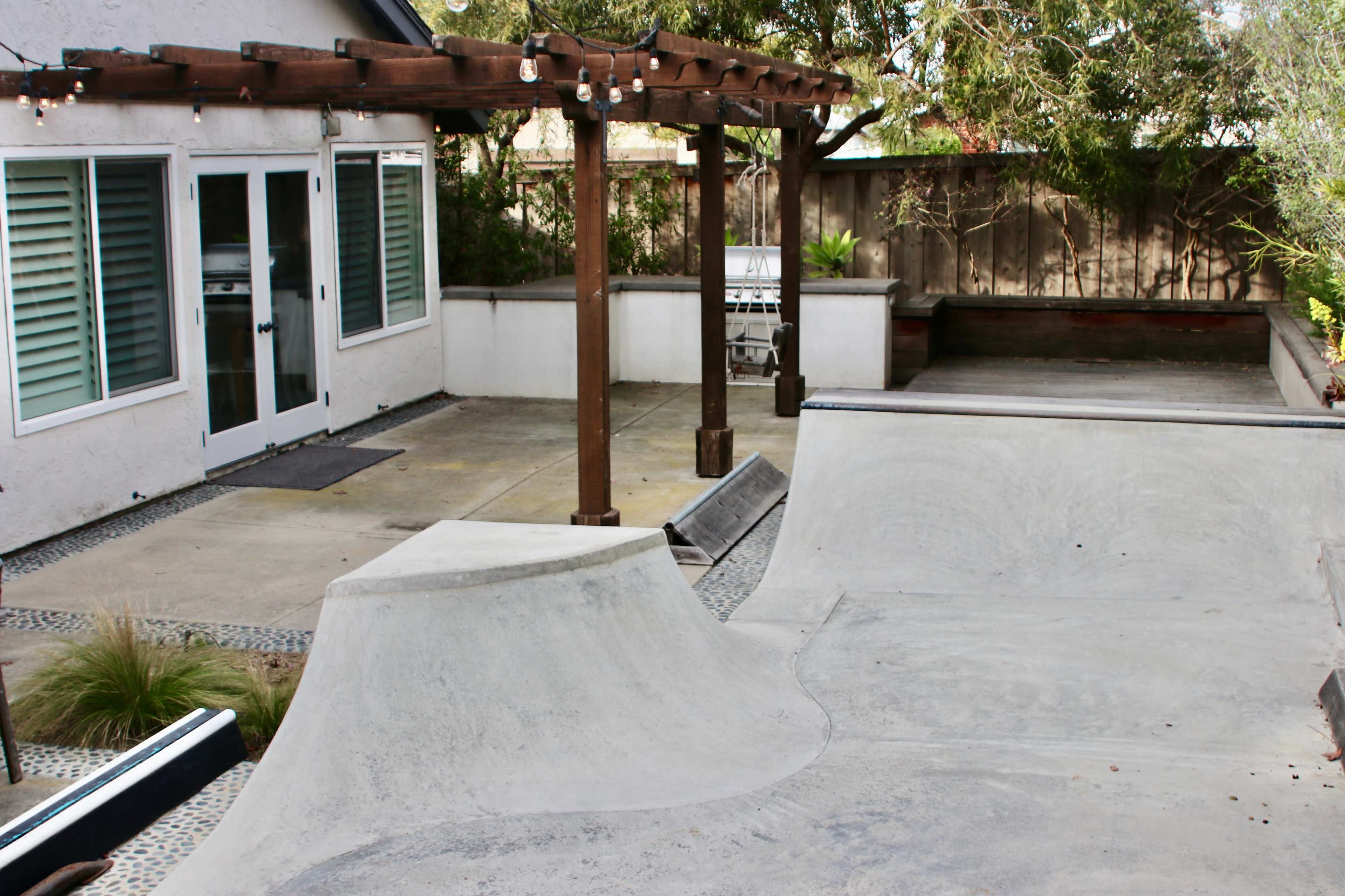 The image shows a backyard skatepark with two concrete ramps and a patio area featuring a wooden pergola, outdoor grill, and greenery.