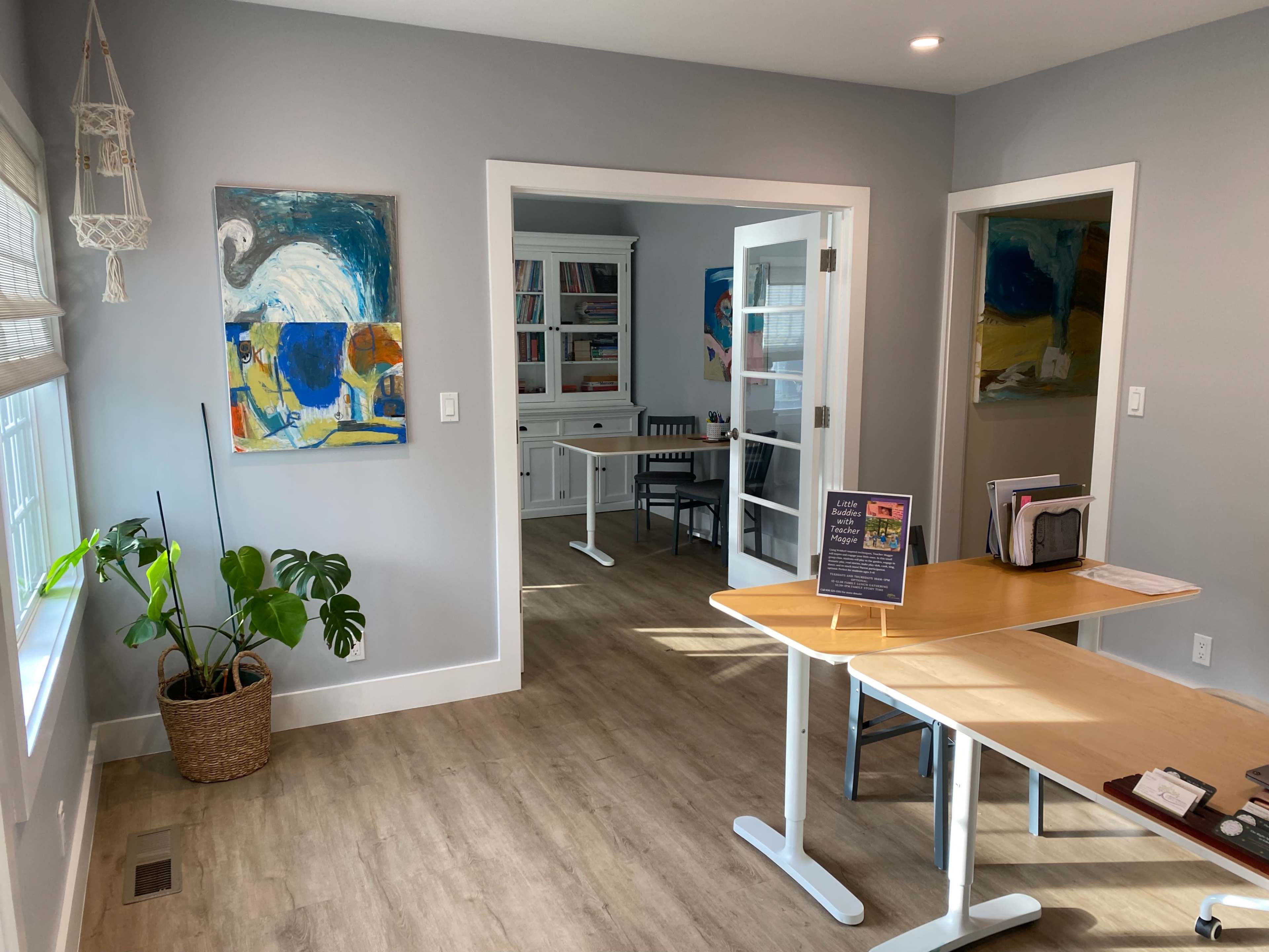 A modern office space with light gray walls, a wooden table, and artwork on the walls, featuring a potted plant in the corner and an adjoining room visible through glass doors.