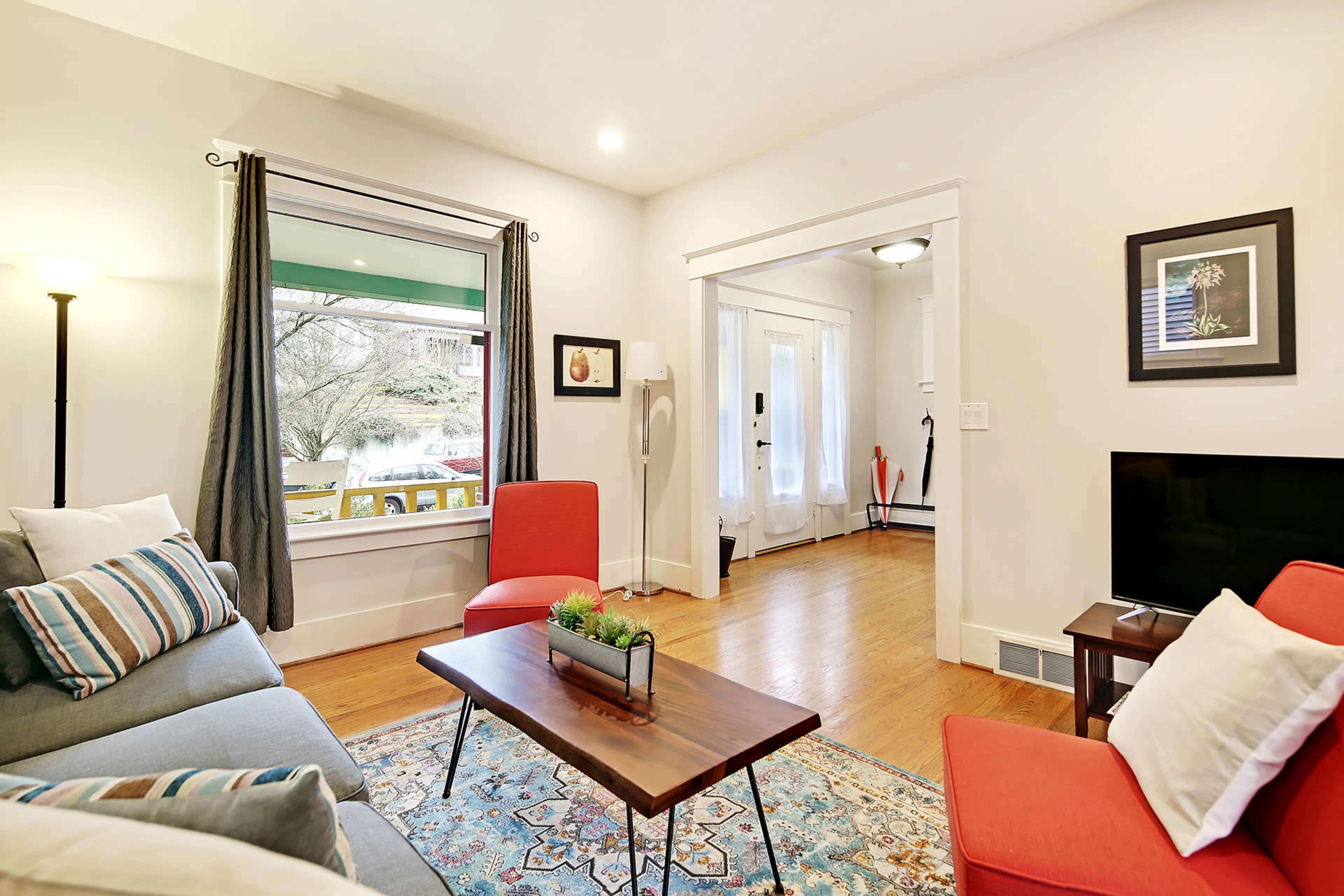 A living room features a gray sofa with colorful throw pillows, a wooden coffee table, and red chairs, with a view of an adjacent room through an open doorway.