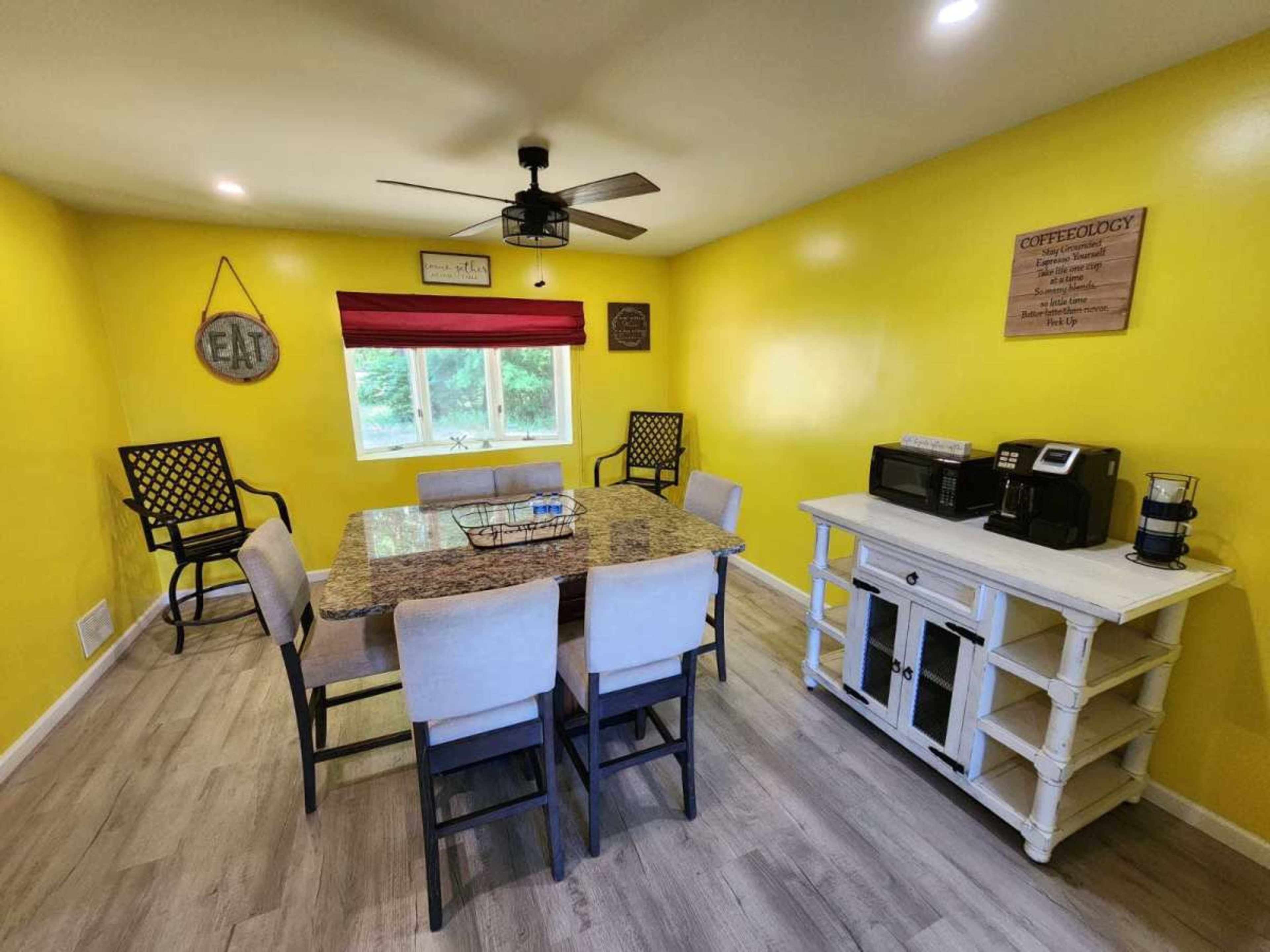 The image shows a dining area with a large table surrounded by chairs, yellow walls, a ceiling fan, and a coffee station on a white sideboard.