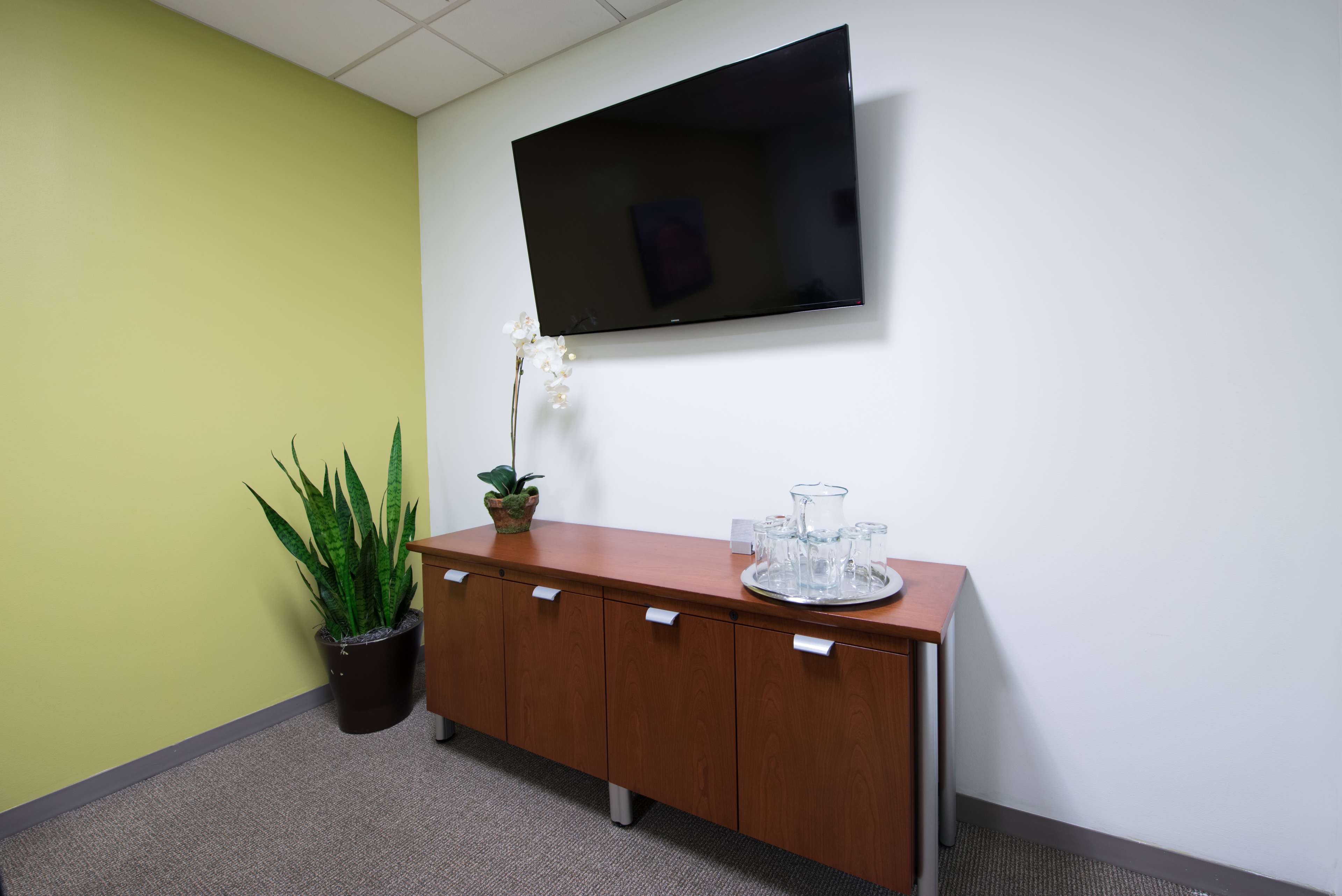 An office corner with a wooden sideboard, a large flat-screen television mounted on the wall, a decorative plant, and a tray with glasses and a pitcher.