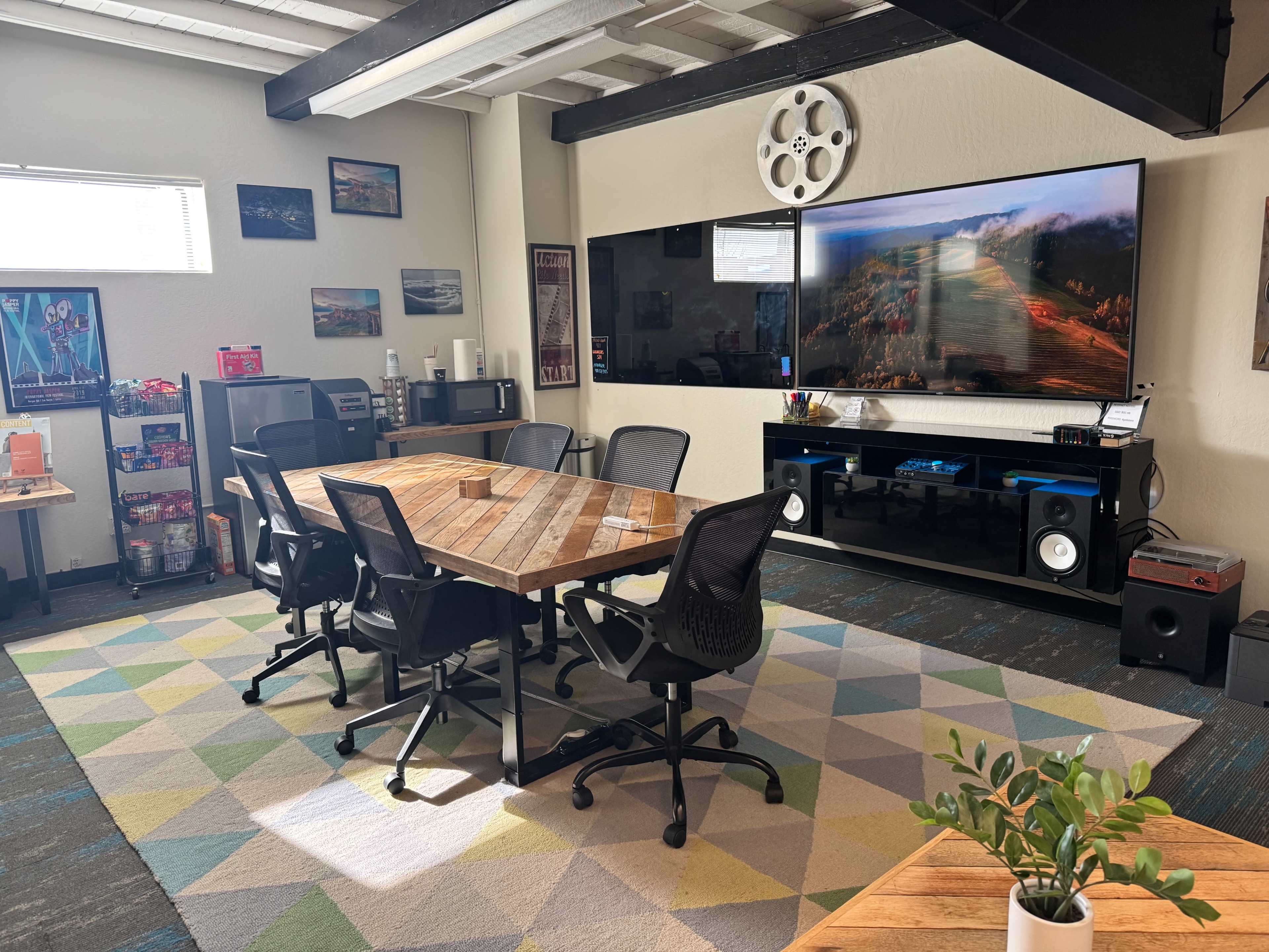 The image shows a modern conference room with a wooden table surrounded by black office chairs, a large wall-mounted television, and various decorative elements and equipment in the background.