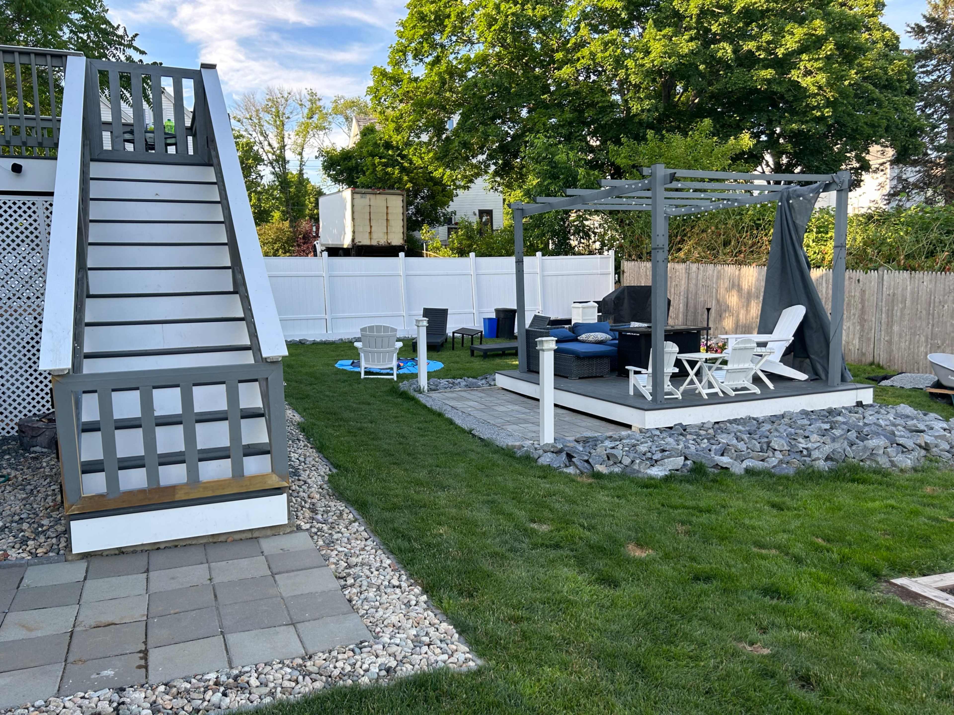 A backyard scene with a set of stairs leading to a platform on the left and a patio area with furniture and a pergola on the right.