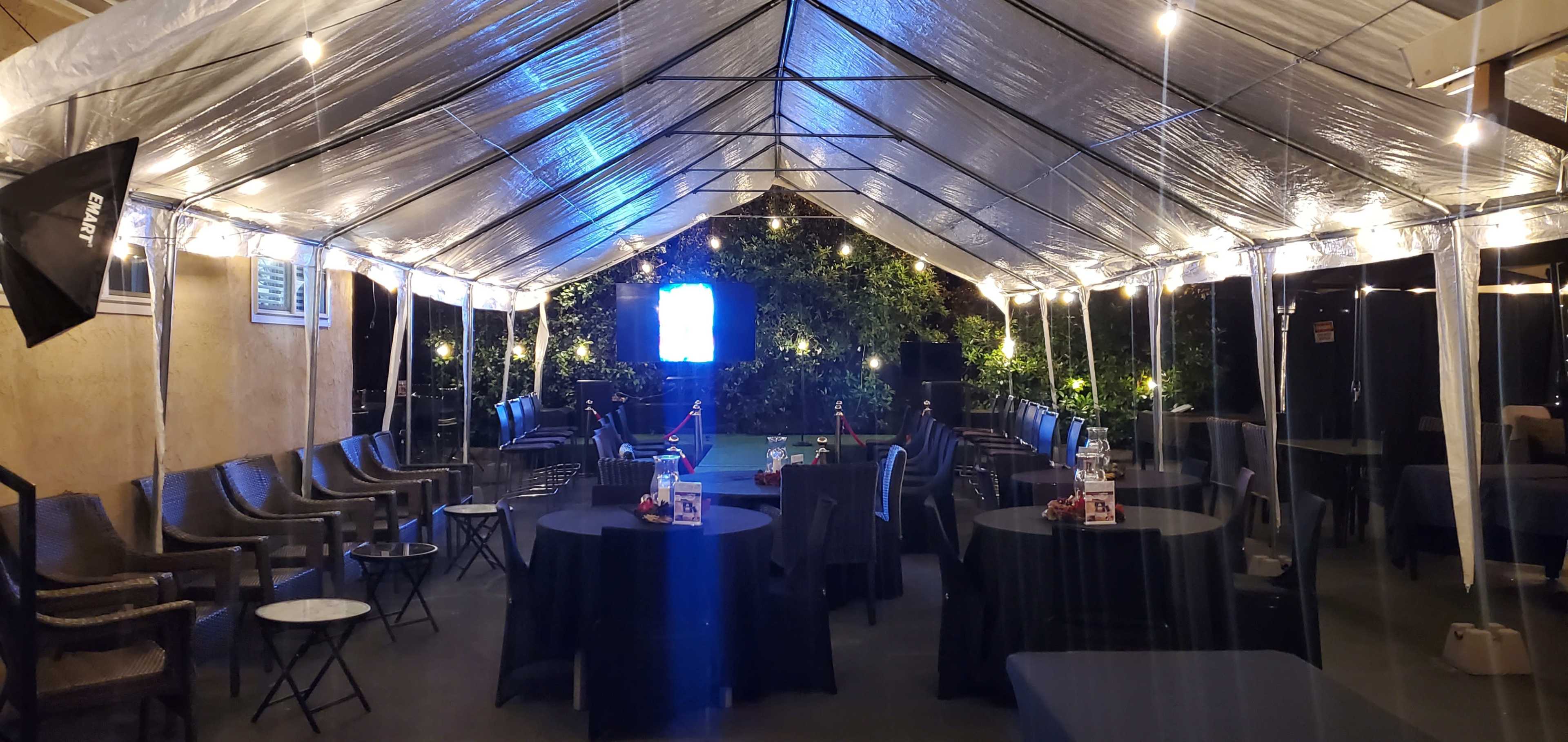 A tented outdoor space is set up with tables and chairs, featuring dim lighting and a large screen at one end, surrounded by greenery.