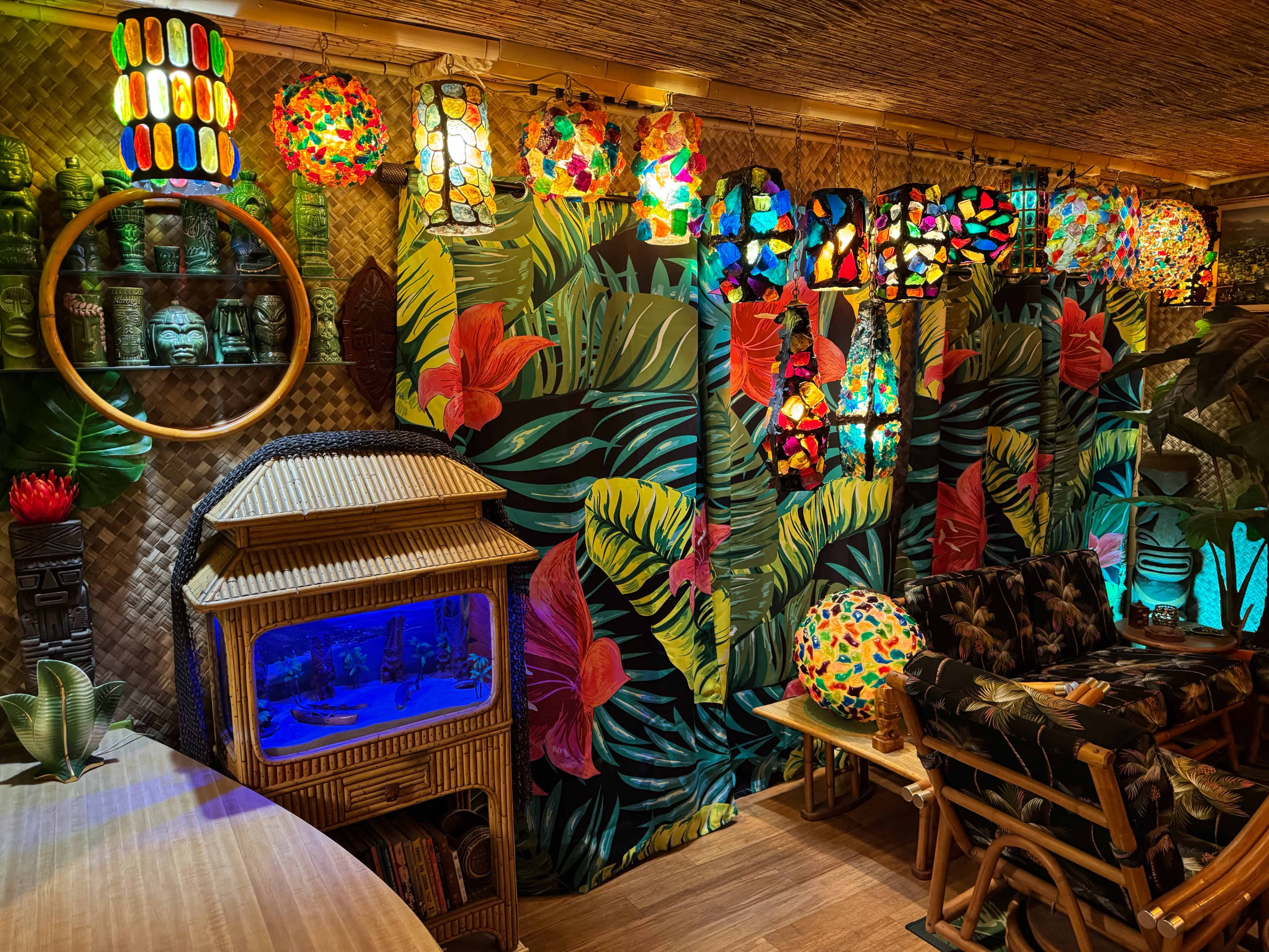 The scene features a brightly decorated room with a tropical theme, showcasing colorful glass lamps, a wall adorned with large green leaves and red flowers, and a bamboo-style aquarium unit.