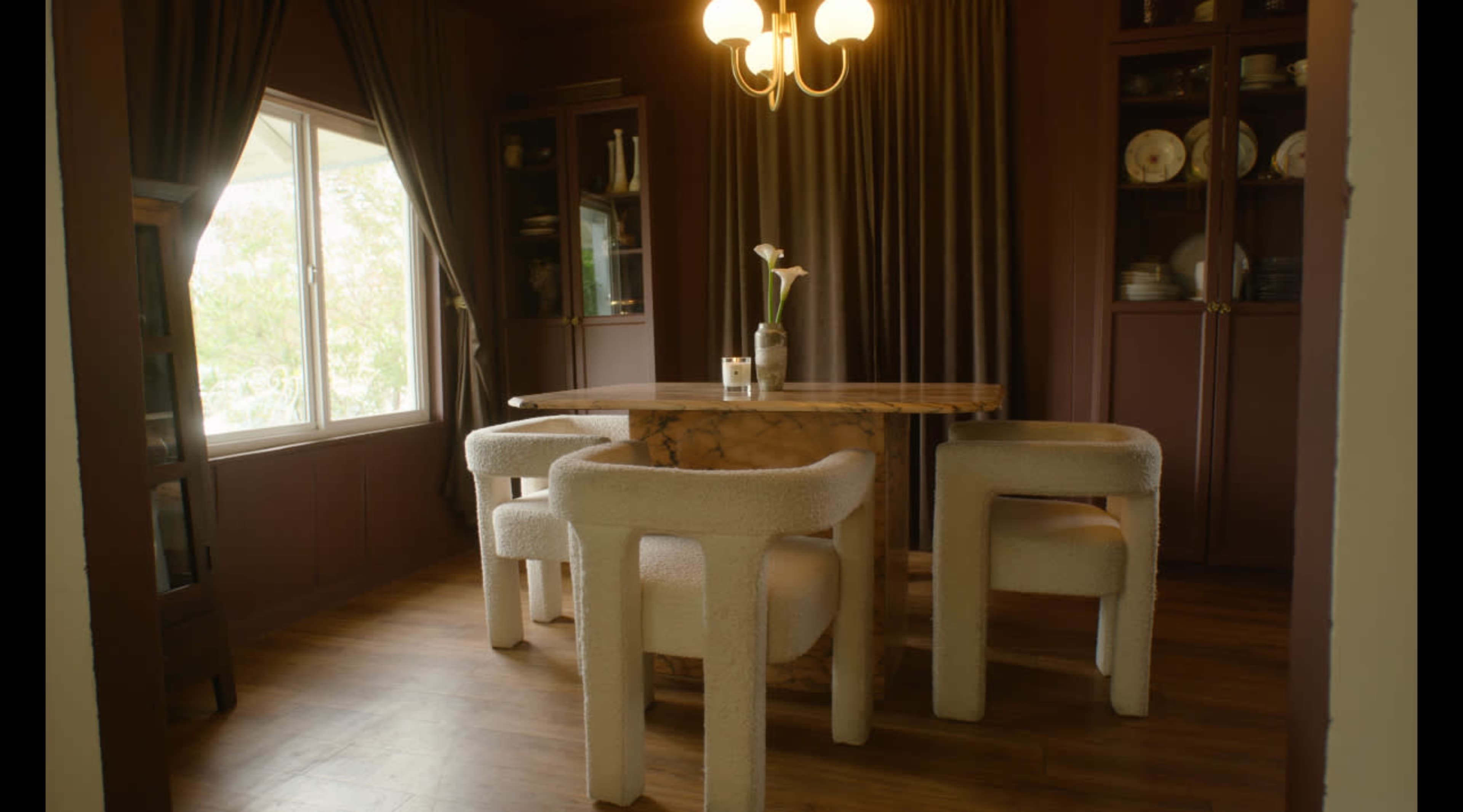 A dining area features a round, marbled table surrounded by four upholstered chairs, with large windows and dark curtains in the background.