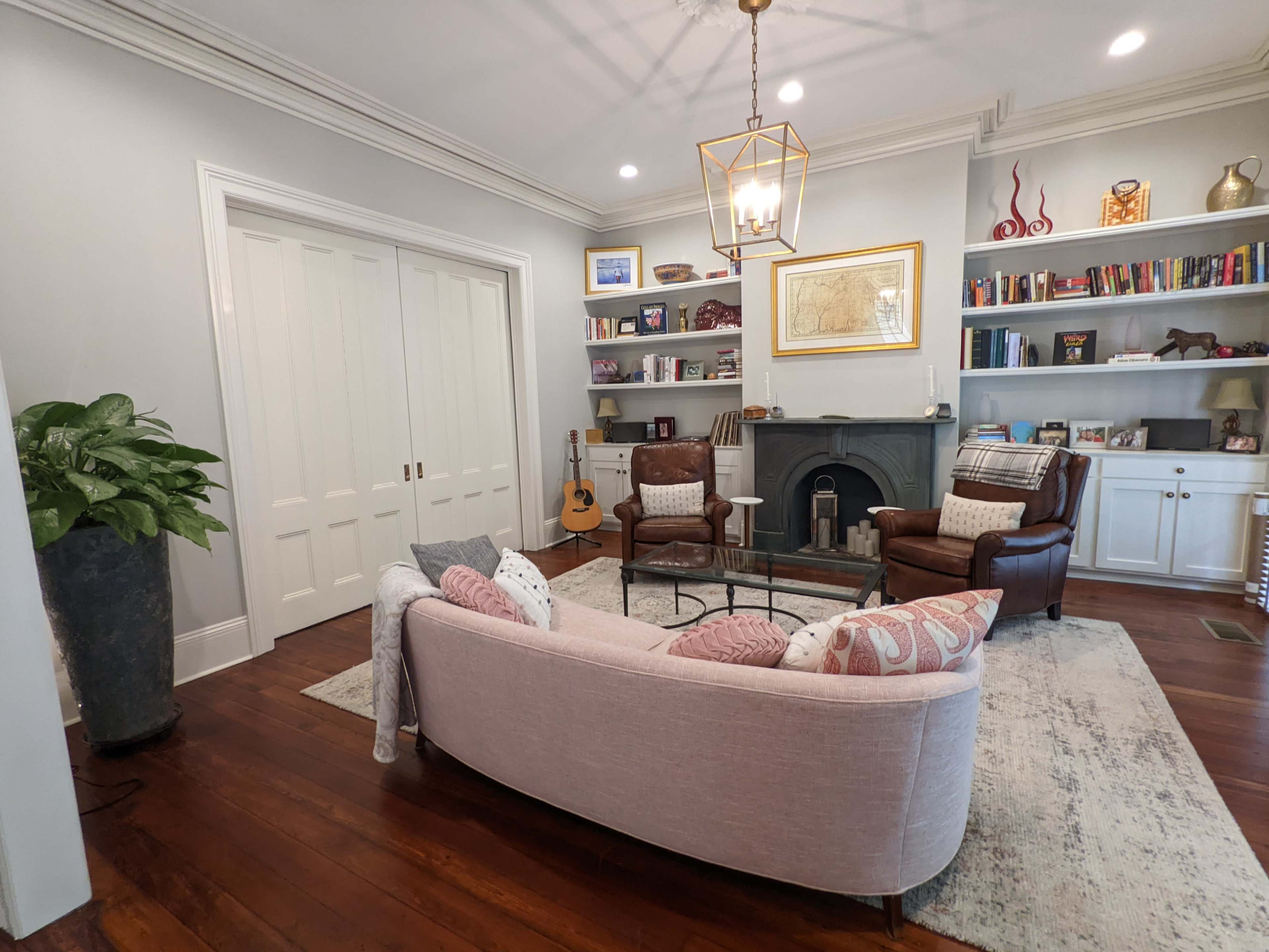 A cozy living room features a pink sectional sofa, two brown armchairs, a glass coffee table, and a bookshelf filled with books, alongside a guitar leaning against the wall.