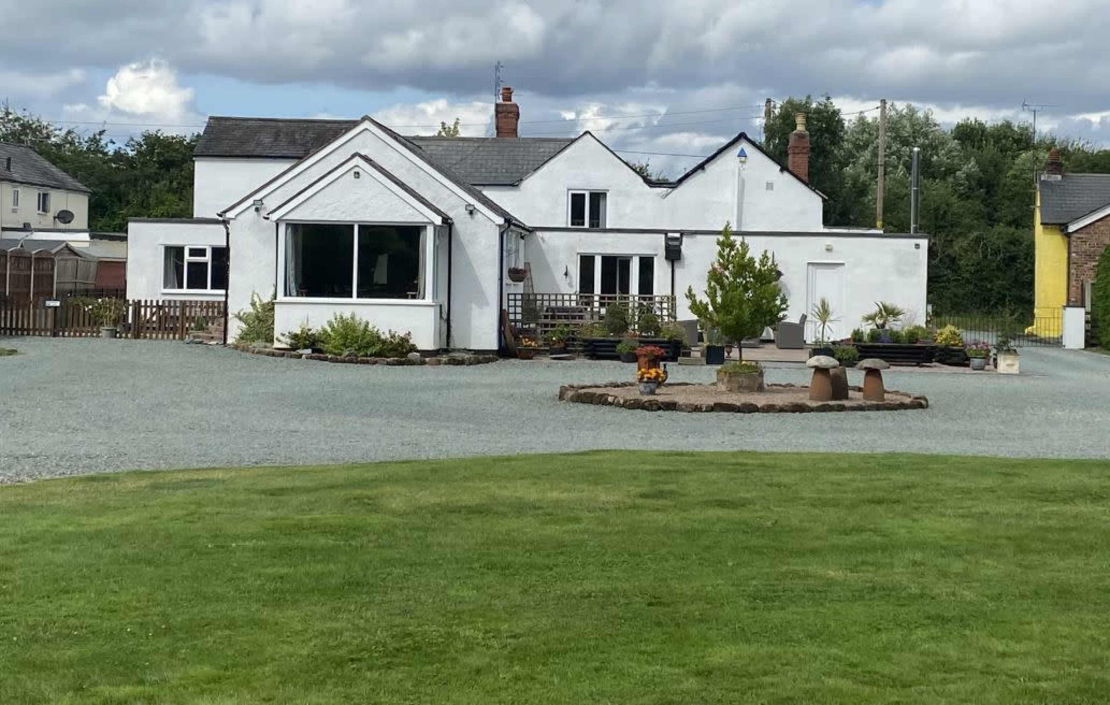 A white, single-story house with a large front window is set on a gravel lot surrounded by a green lawn and landscaped flower beds.