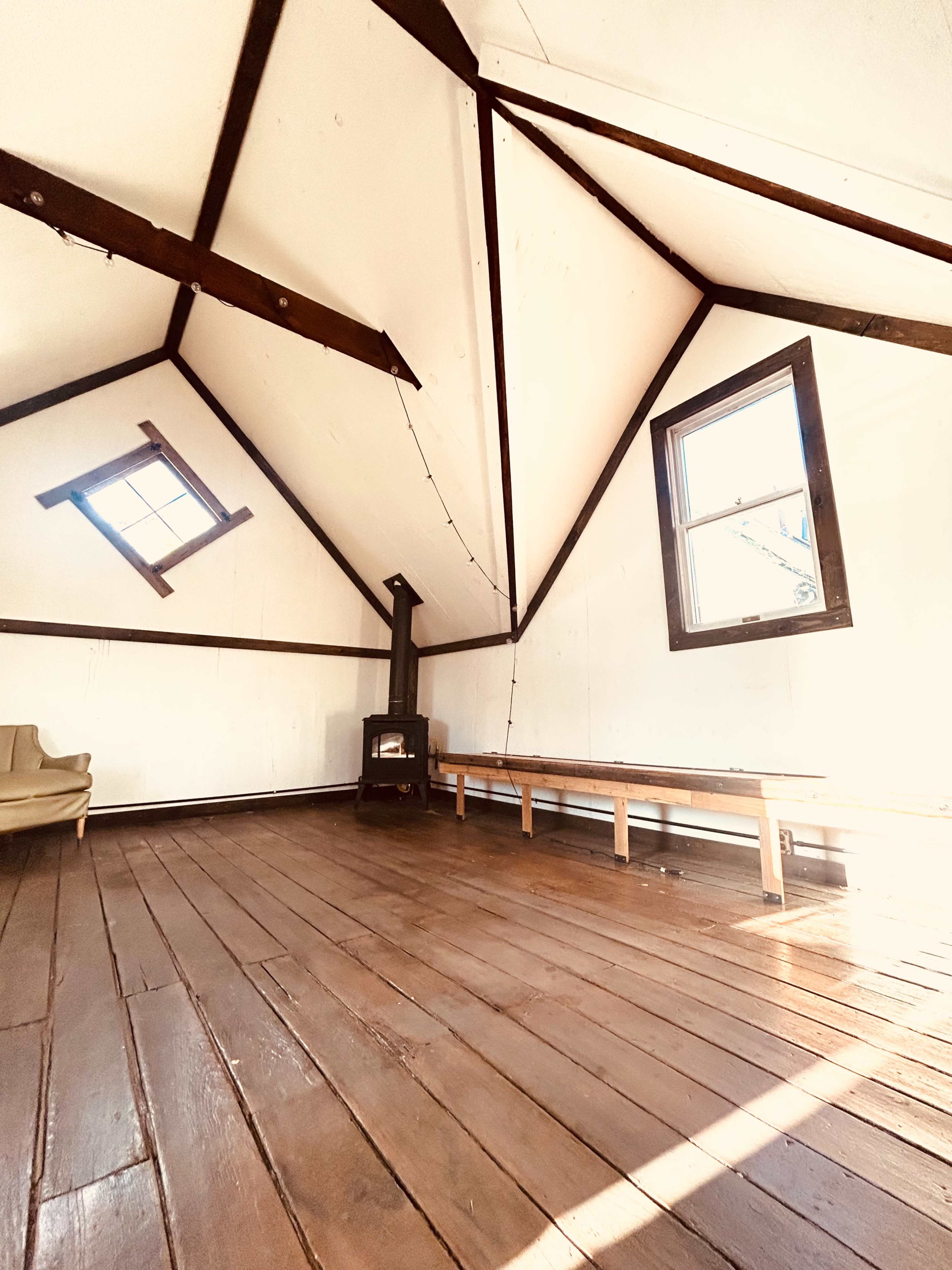 The image shows an empty room with a high, beamed ceiling, wooden floors, a small stove, a long bench, and a window letting in natural light.