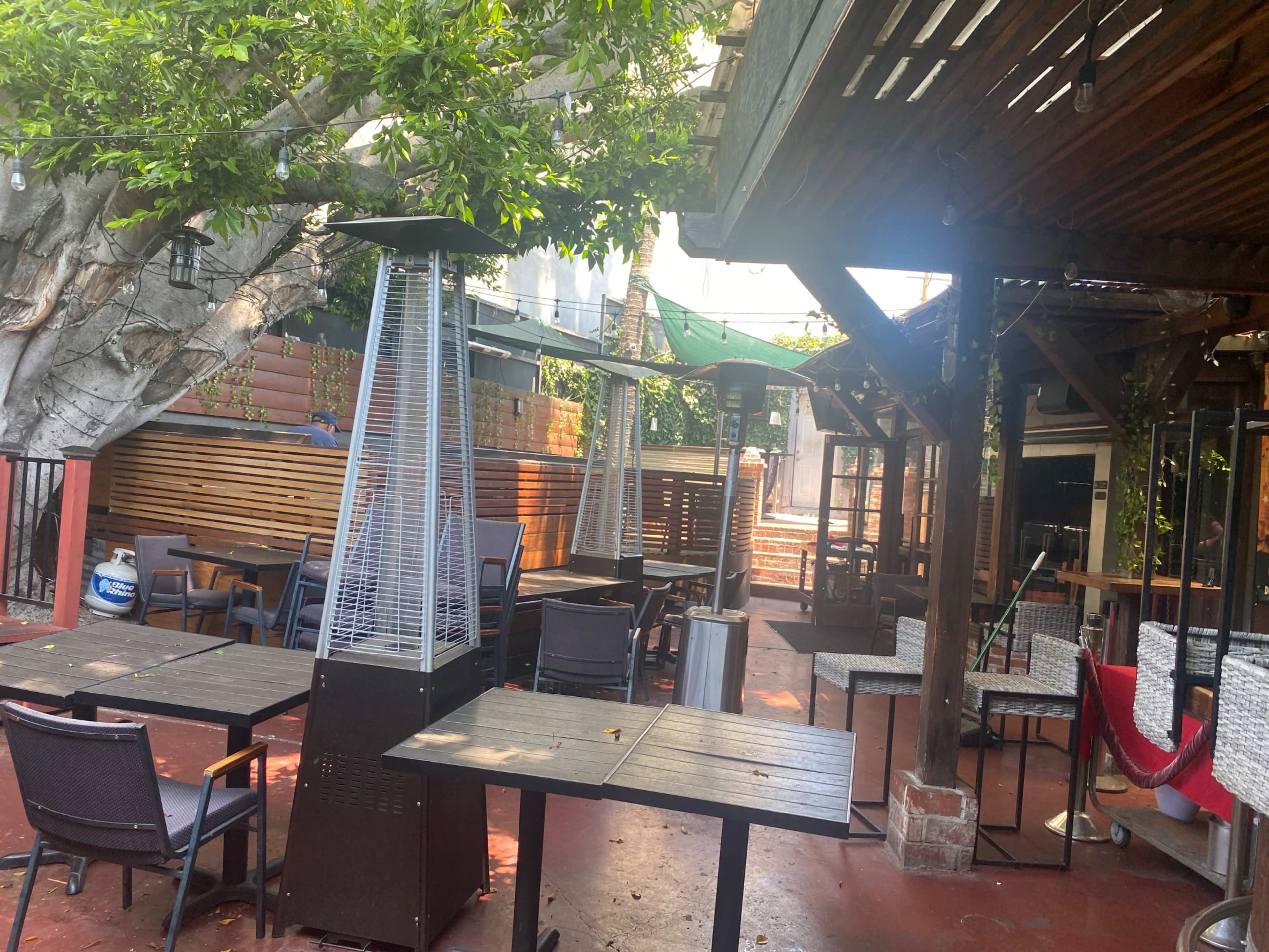 The image shows an outdoor dining area with several tables, heaters, and surrounding greenery.