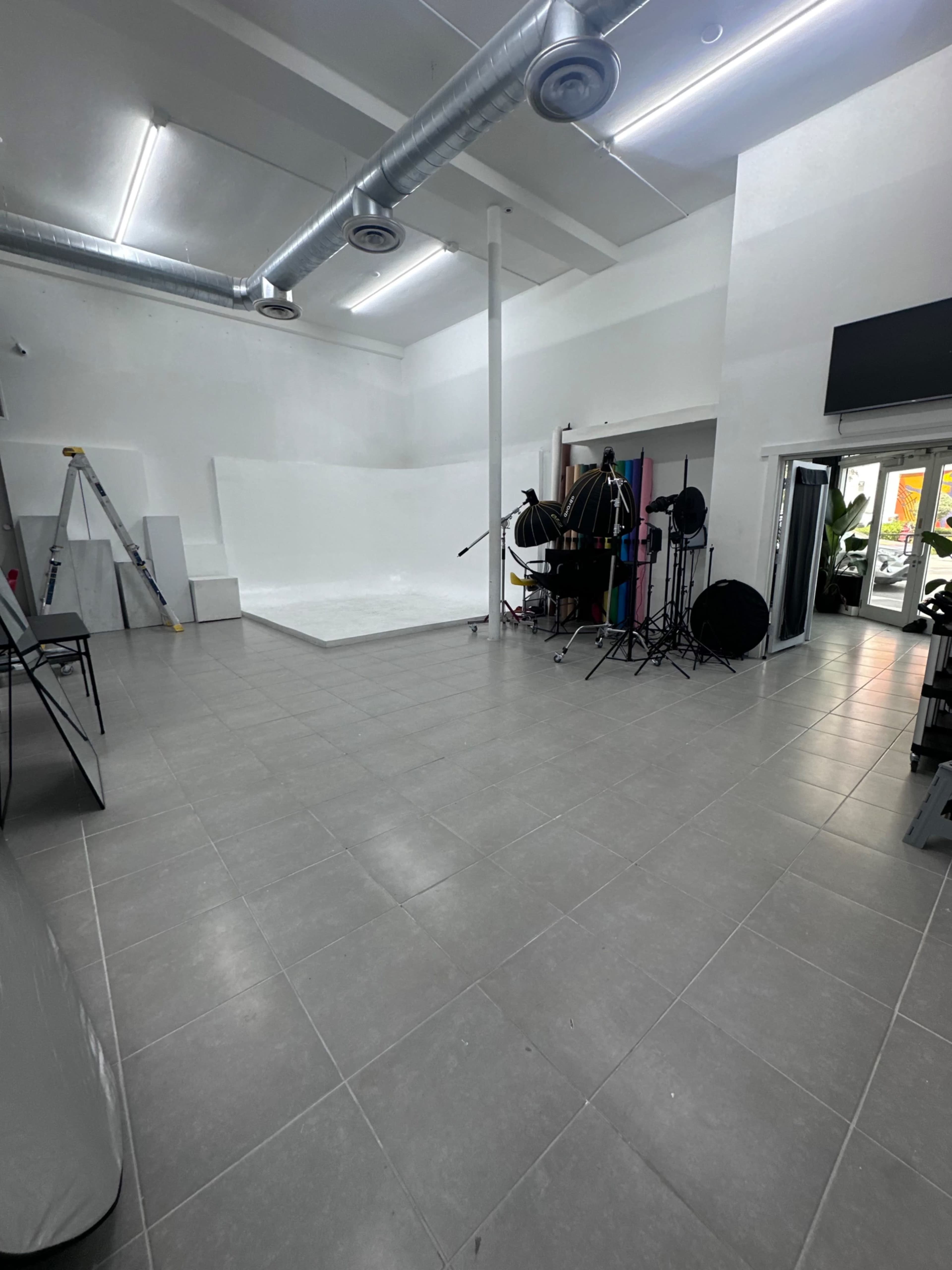 The image shows a spacious photography studio with a white backdrop, lighting equipment, a ladder, and gray tiled flooring.