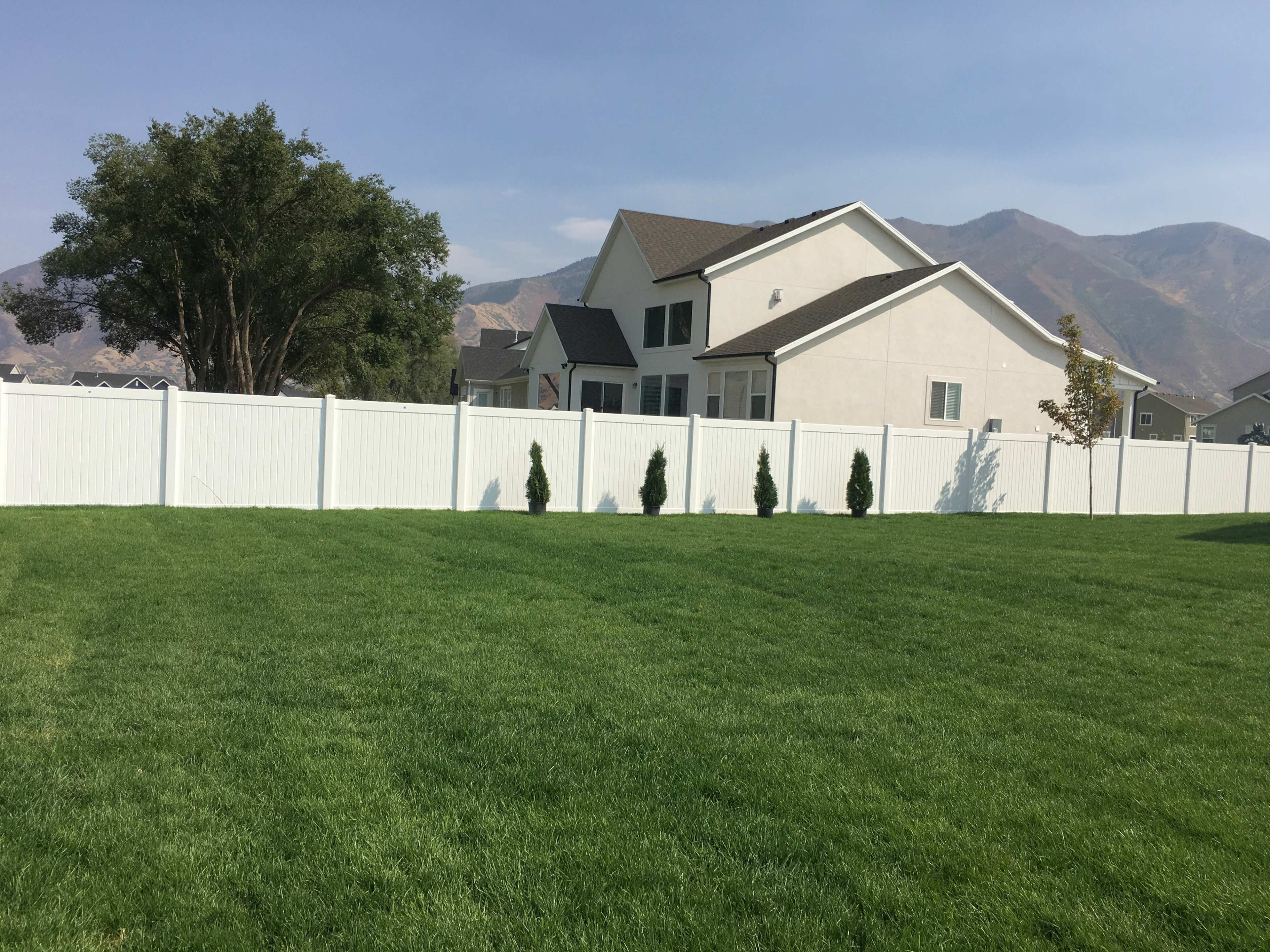 A white fence surrounds a well-maintained green lawn in front of a two-story house with a mountain backdrop.