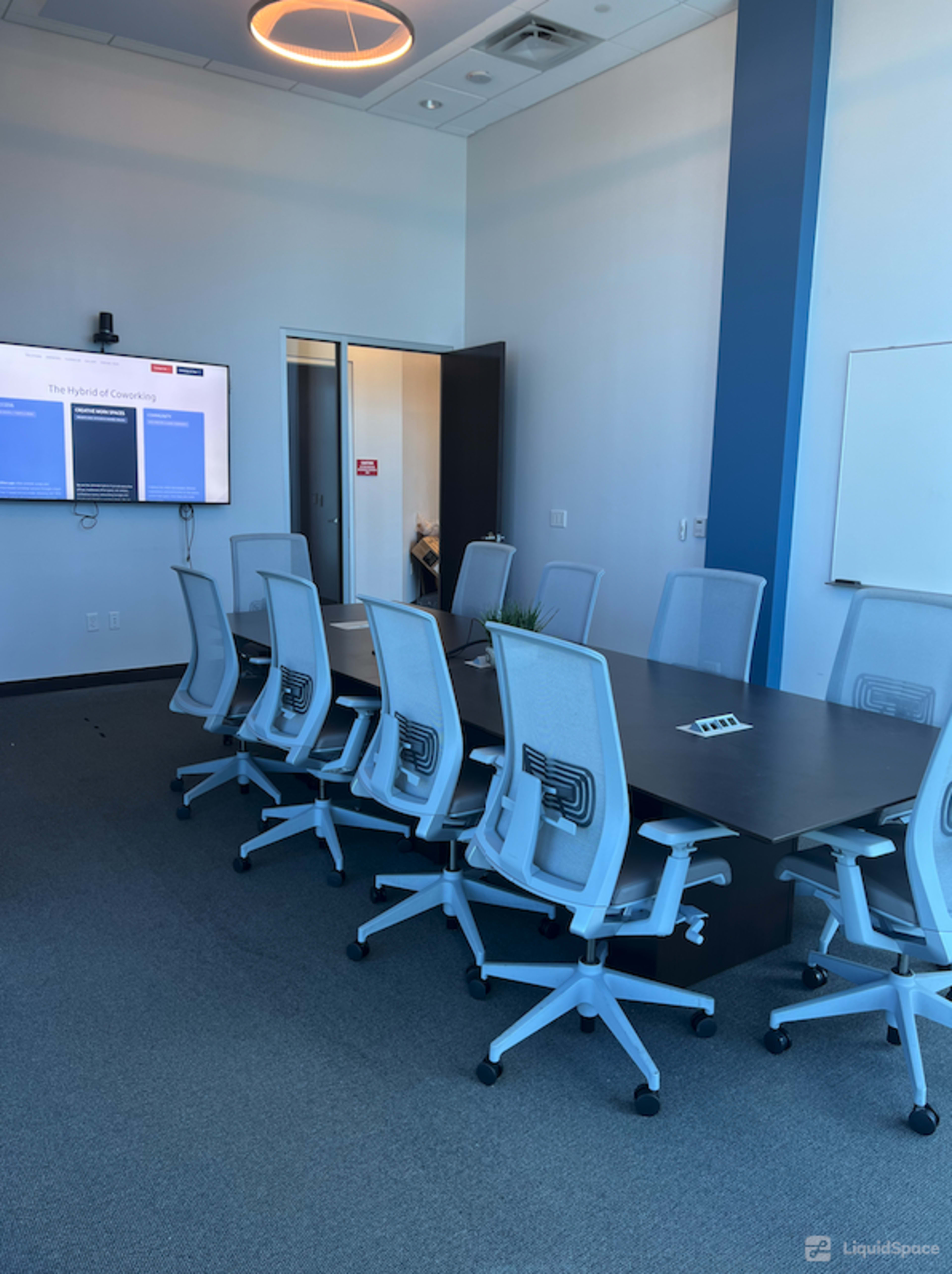 A modern conference room features a long table surrounded by ergonomic chairs, with a large screen displaying content on one wall.