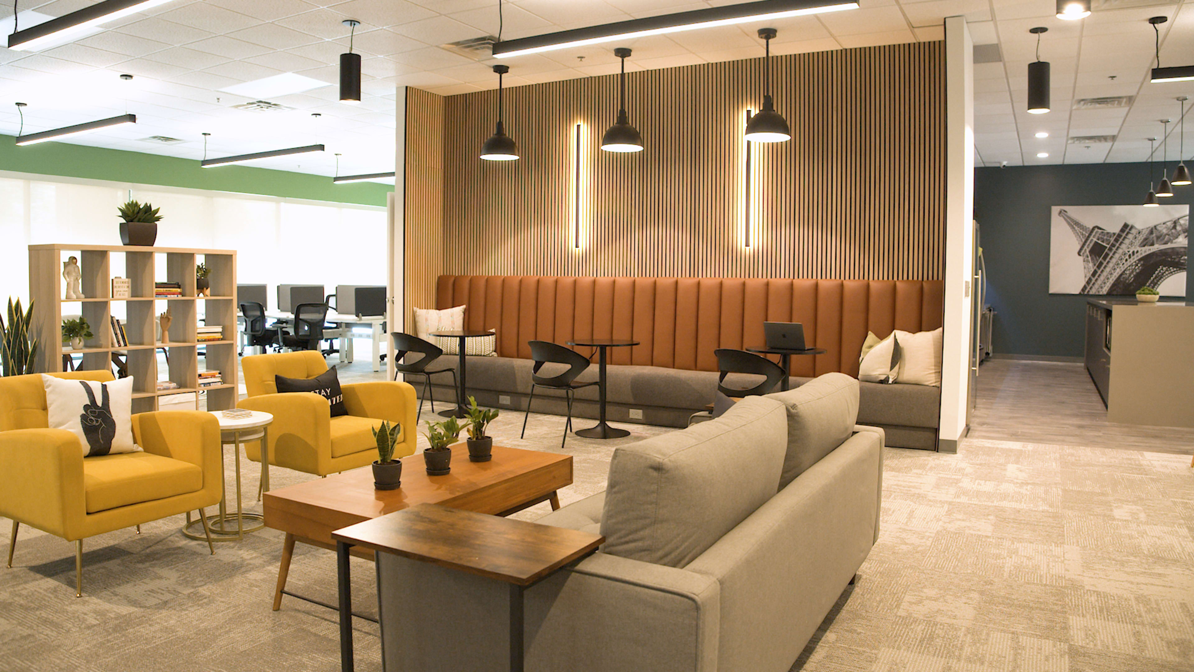 A modern open office space with a mix of seating areas, including yellow chairs and gray sofas, against a backdrop of wood paneling and pendant lights.