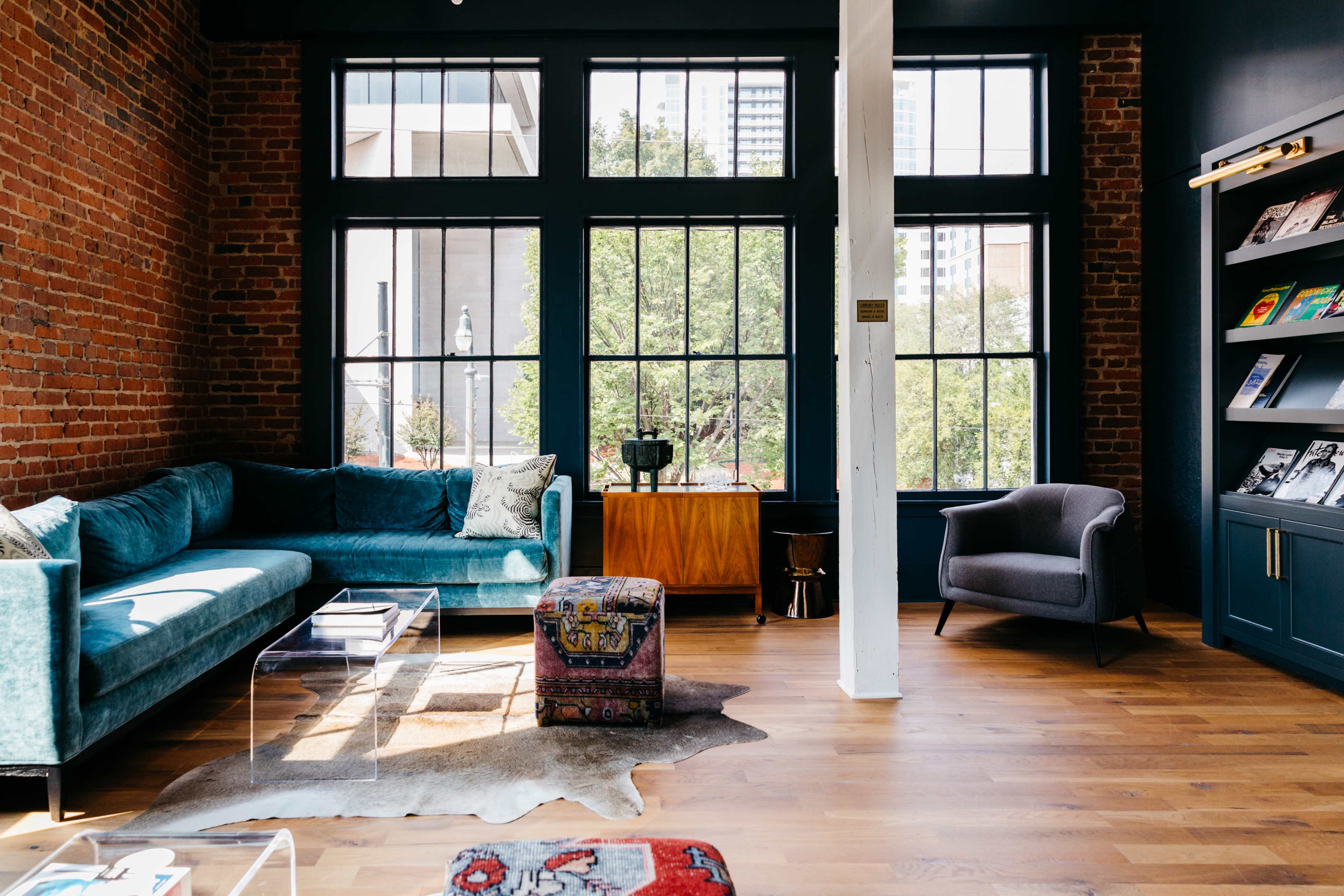 The image shows a cozy living space featuring a teal sofa, a glass coffee table, a colorful pouf, and large windows that provide a view of green trees outside.