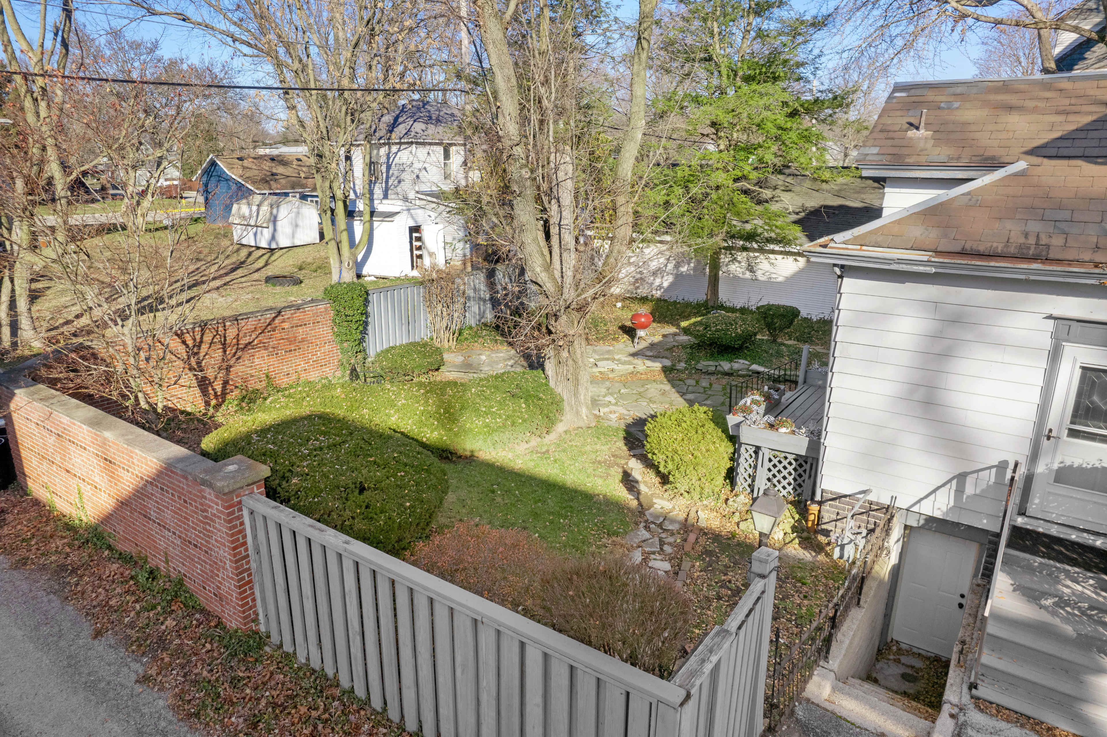 The image shows a residential backyard with a stone path, well-maintained shrubs, and a small wooden deck next to a house.
