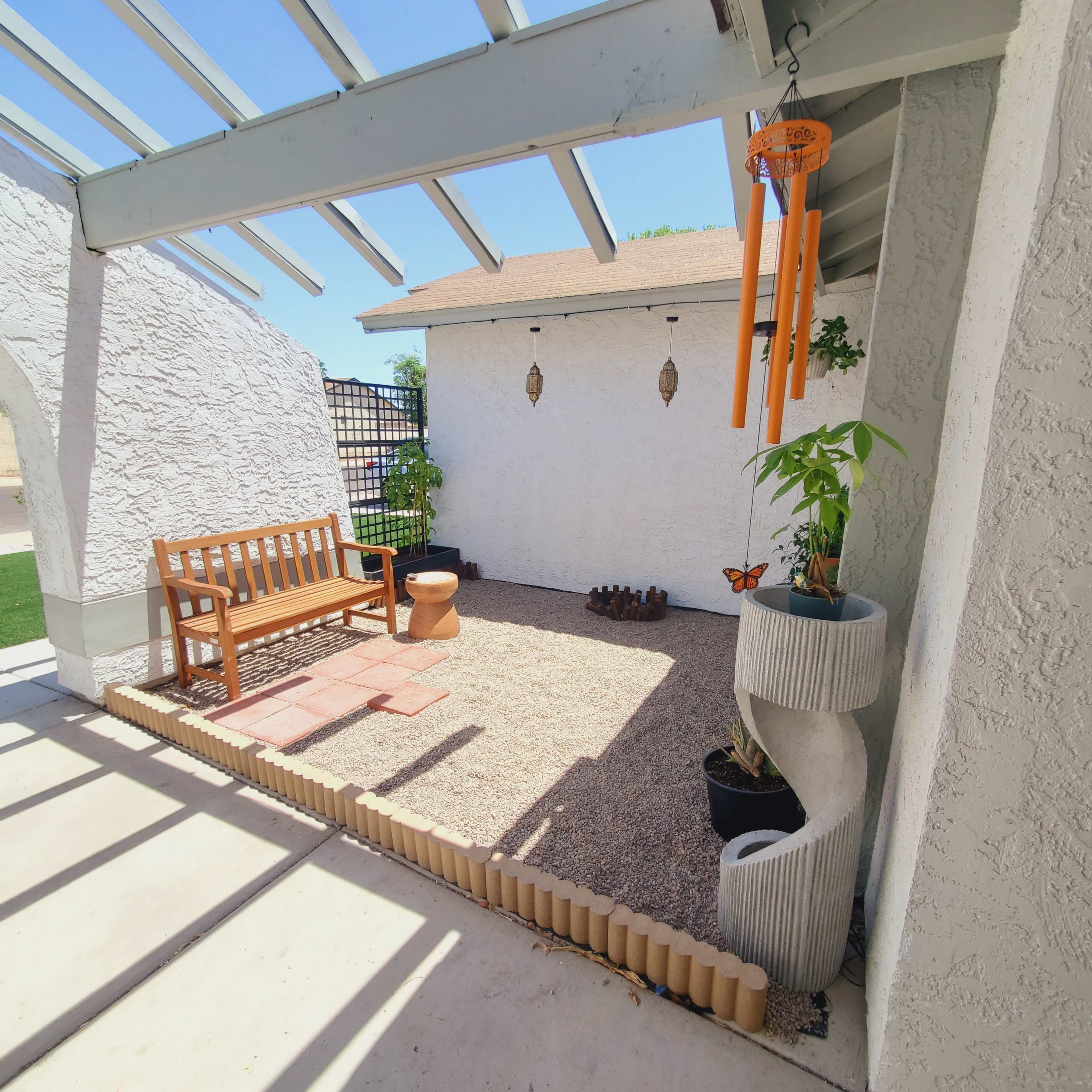 The image shows a small outdoor patio area featuring a wooden bench, a gravel surface, a potted plant, and wind chimes hanging from the ceiling.
