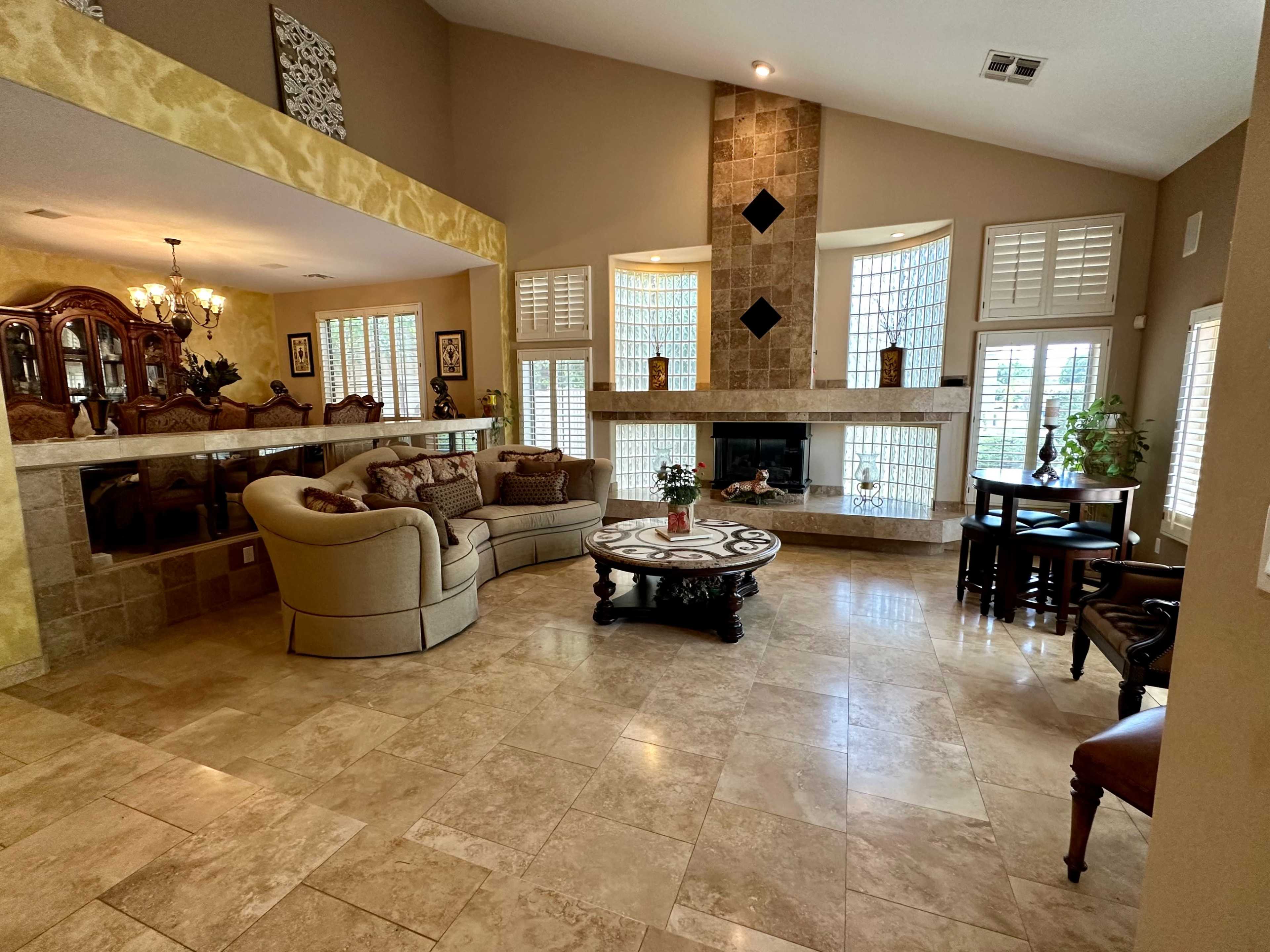The spacious living room features a large beige sectional sofa, a round coffee table, and a fireplace surrounded by stone, with light streaming in through several windows.