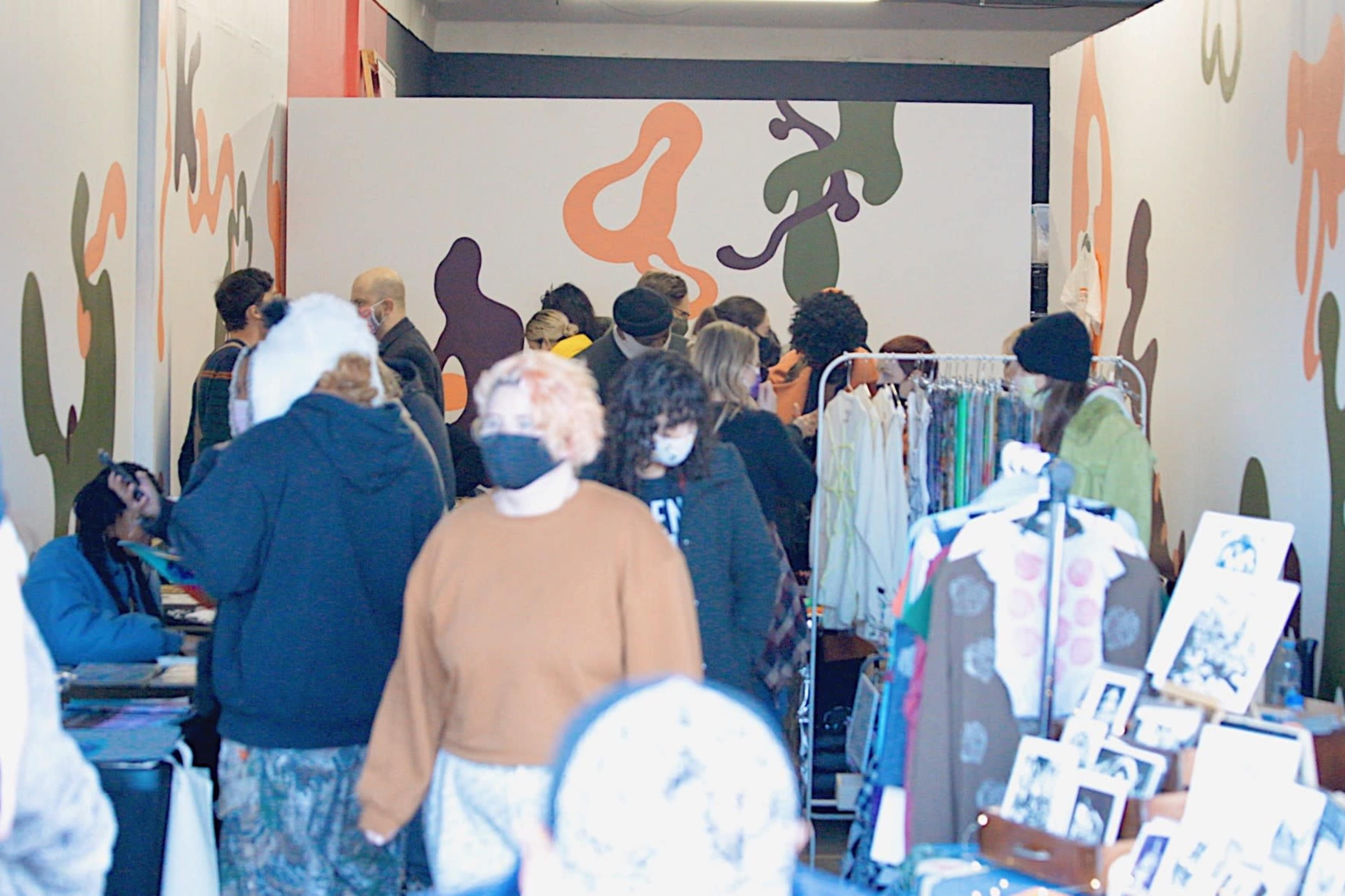 A crowd of people is walking through a narrow space with colorful abstract wall art and various displays of clothing and photographs along the sides.