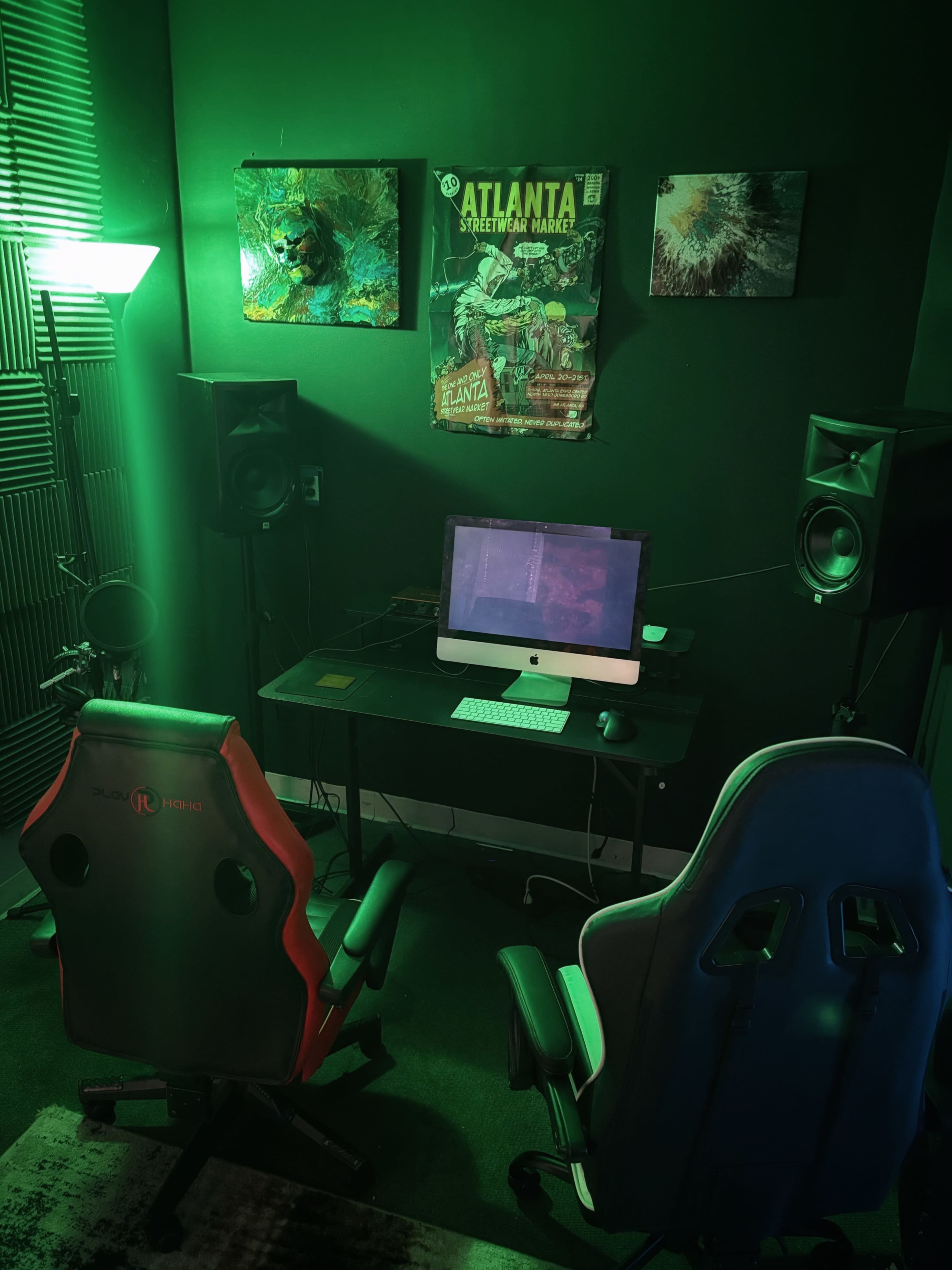 The image shows a gaming setup with two chairs, a desk with a computer, and wall art illuminated by green lighting.