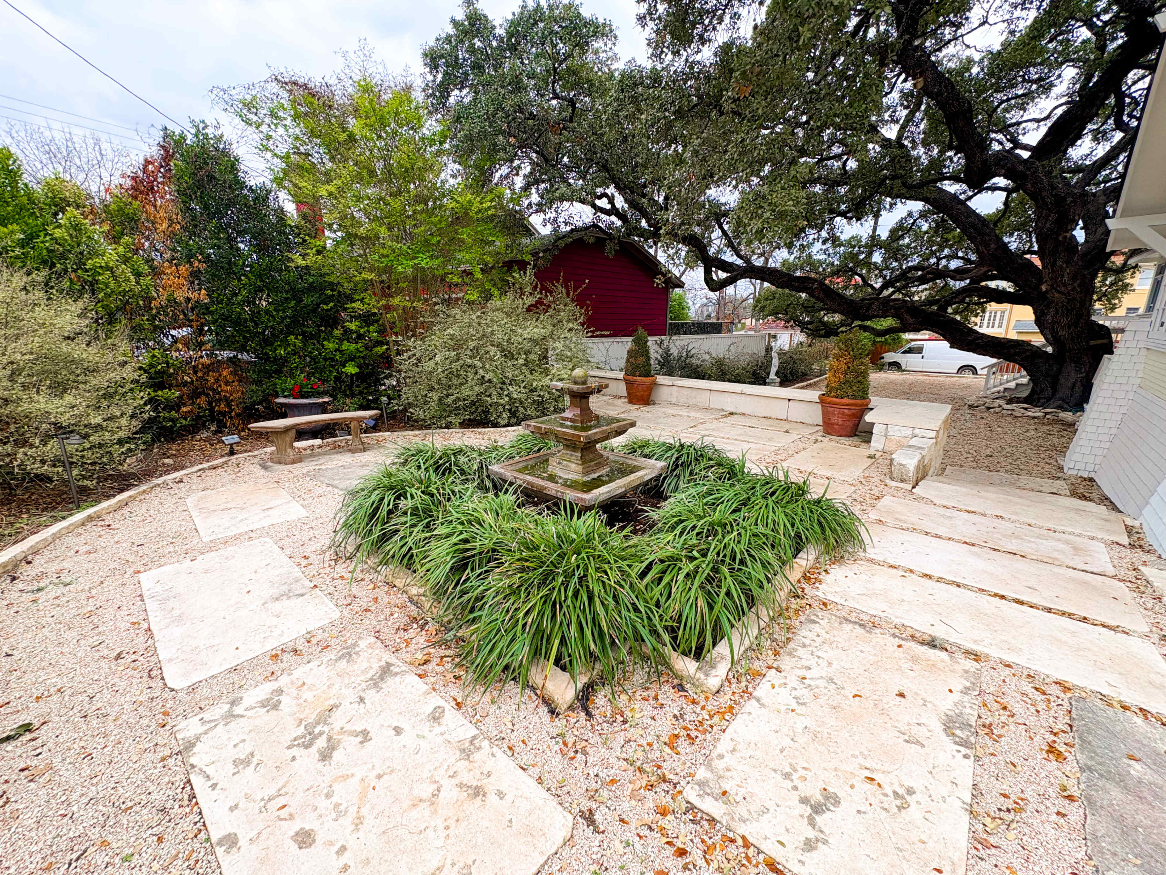 A stone pathway leads to a central fountain surrounded by greenery and decorative pots in a landscaped yard.