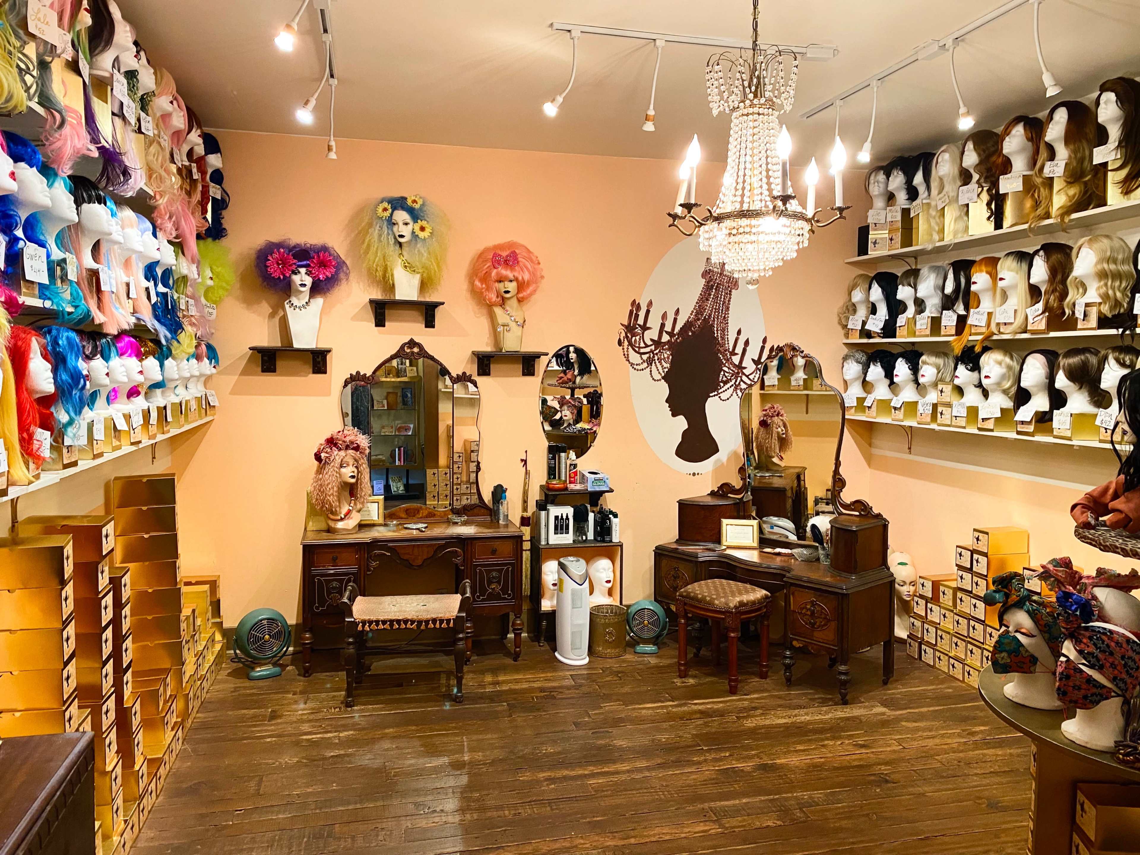The image shows a brightly lit wig shop with an array of colorful wigs displayed on shelves and a vintage-style vanity set in the center.