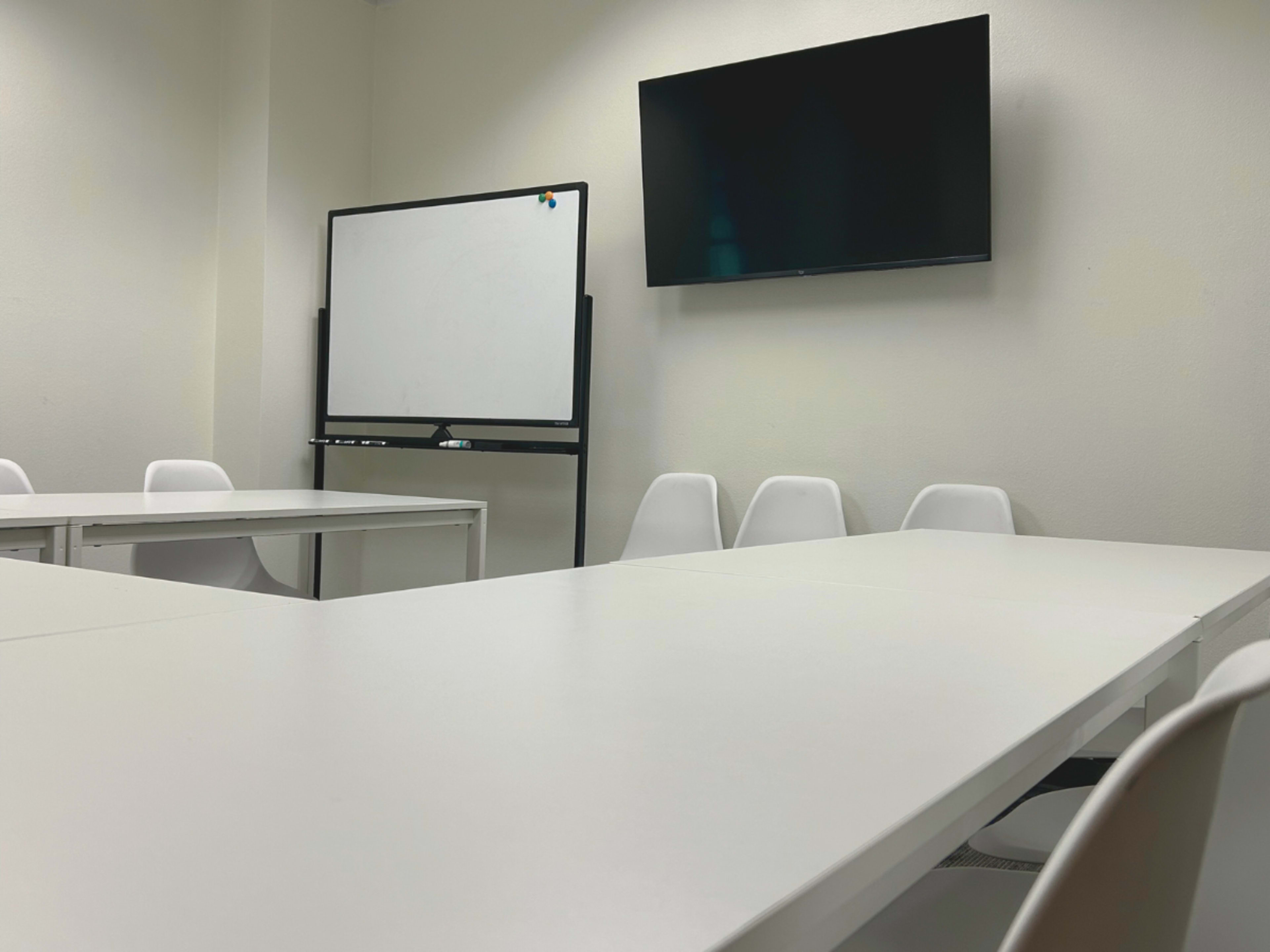 A plain meeting room features a long white table, several white chairs, a whiteboard on a stand, and a wall-mounted television.