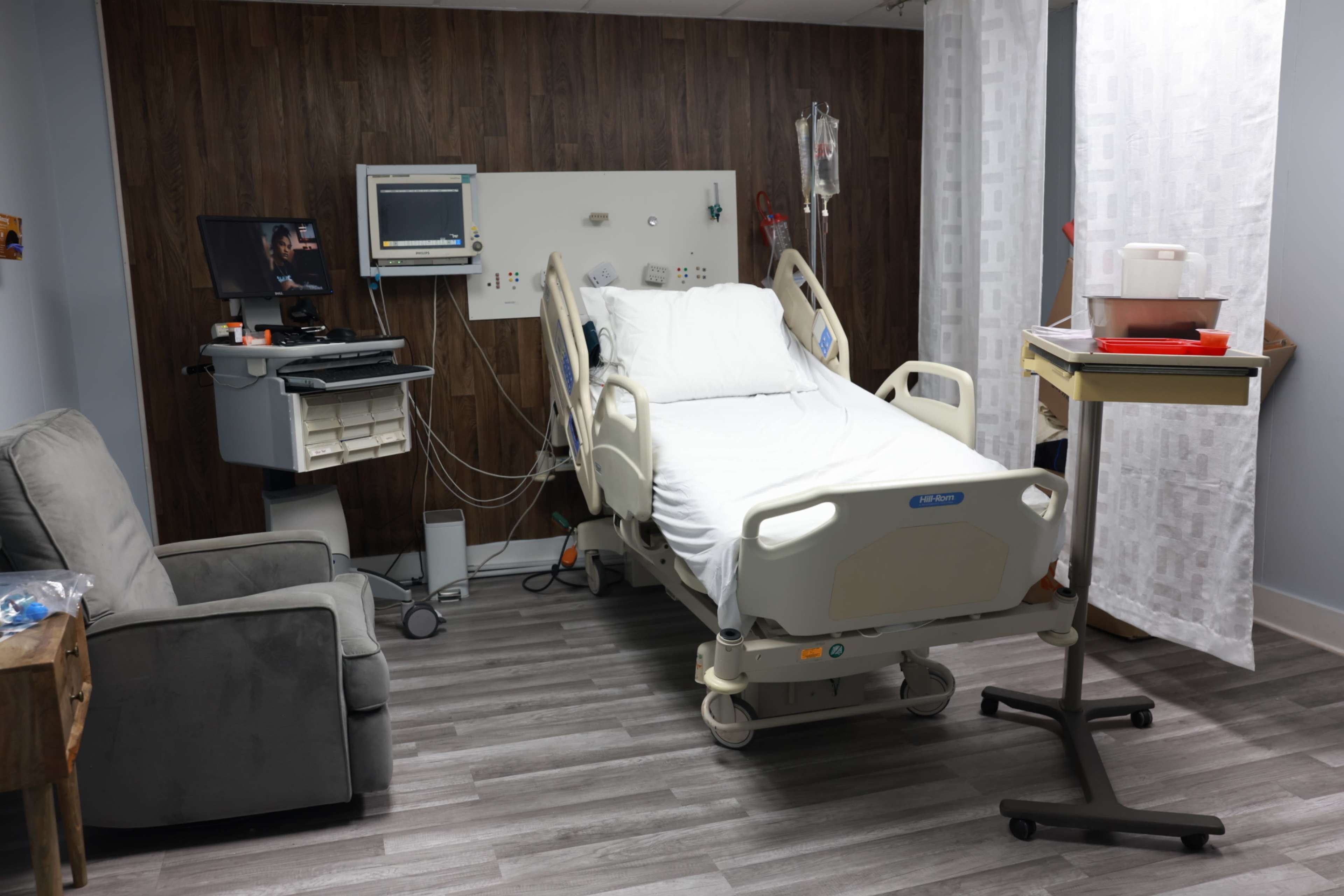 The image shows a hospital room with a patient bed, a medical monitor, and a seating area beside a wooden accent wall.