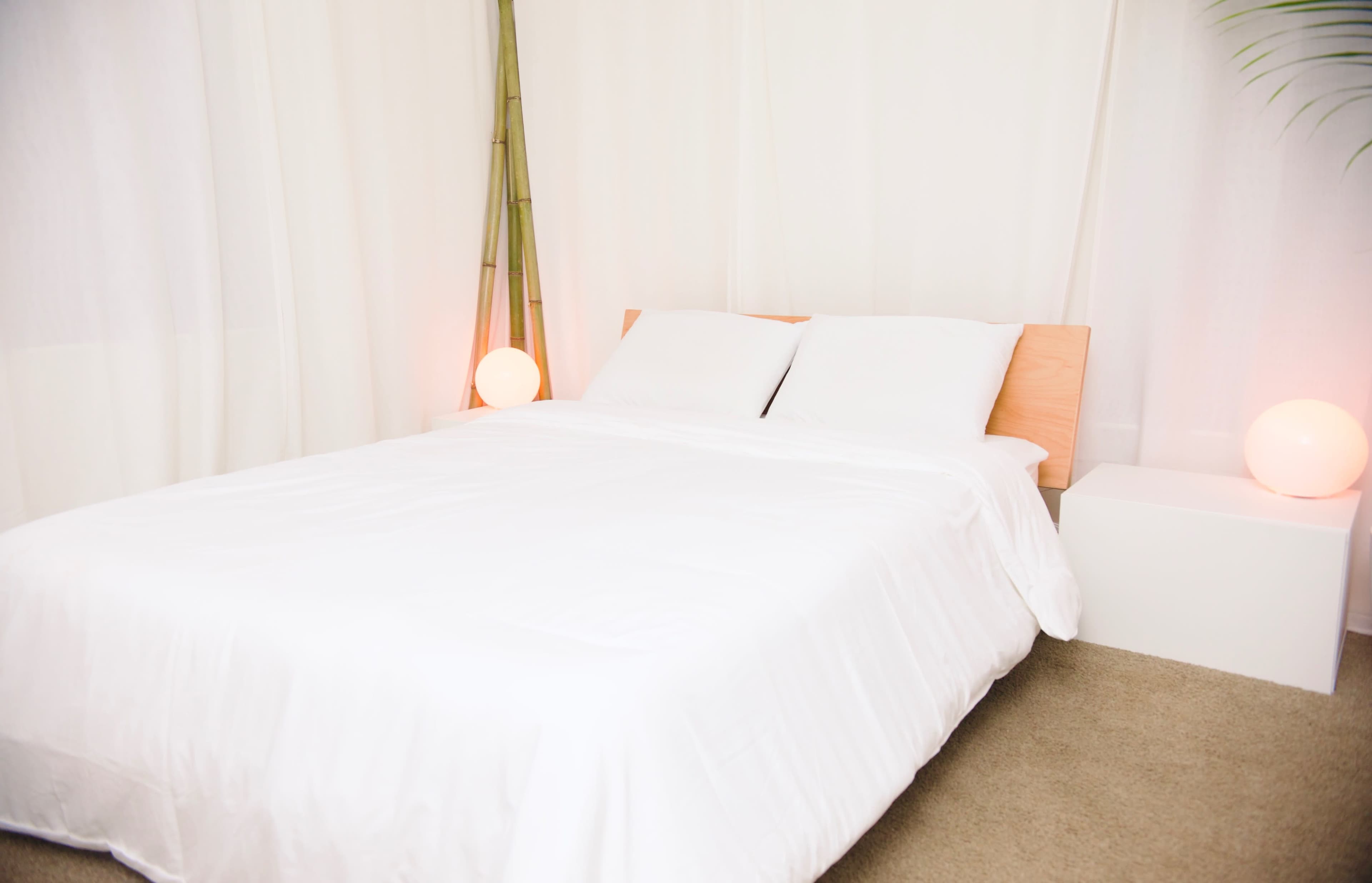 A simple bedroom features a bed with a white comforter and two pillows, accompanied by a side table and soft lighting.