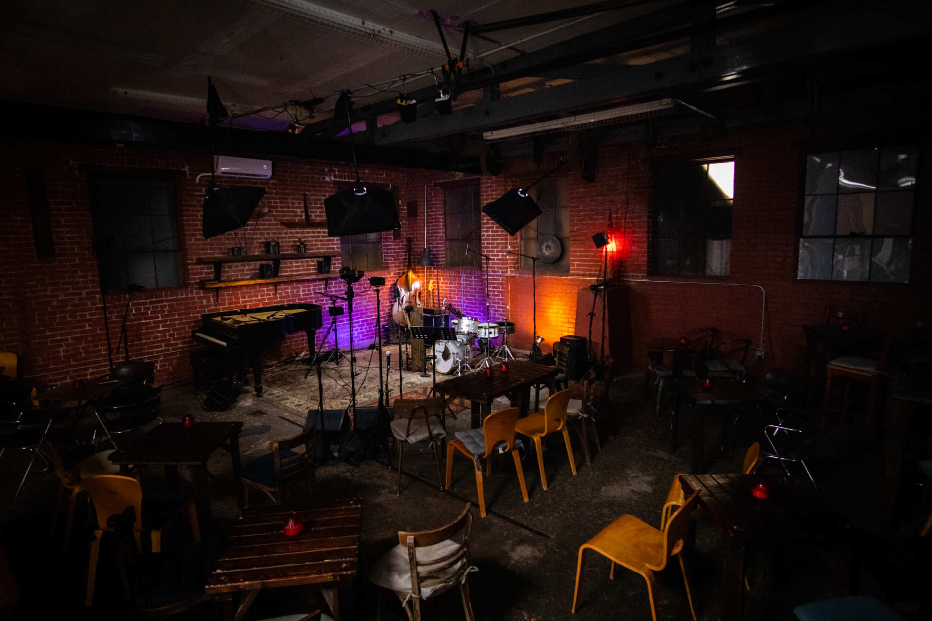 A dimly lit music venue with a stage set up for a performance, featuring a drum kit, microphone, and a piano, surrounded by empty chairs and tables.