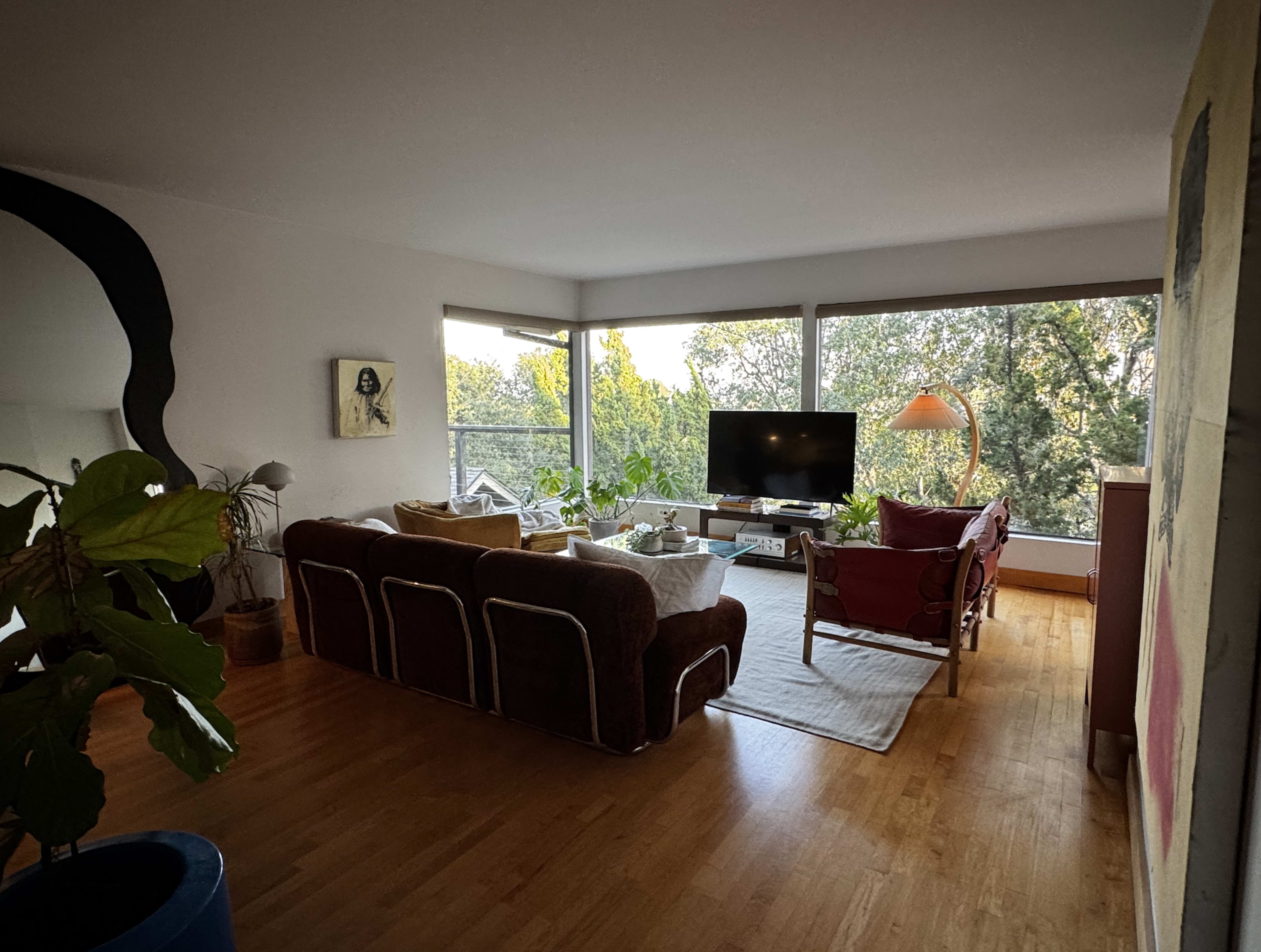The image depicts a modern living room with a brown sofa, red armchairs, a large television, and large windows overlooking a green landscape.