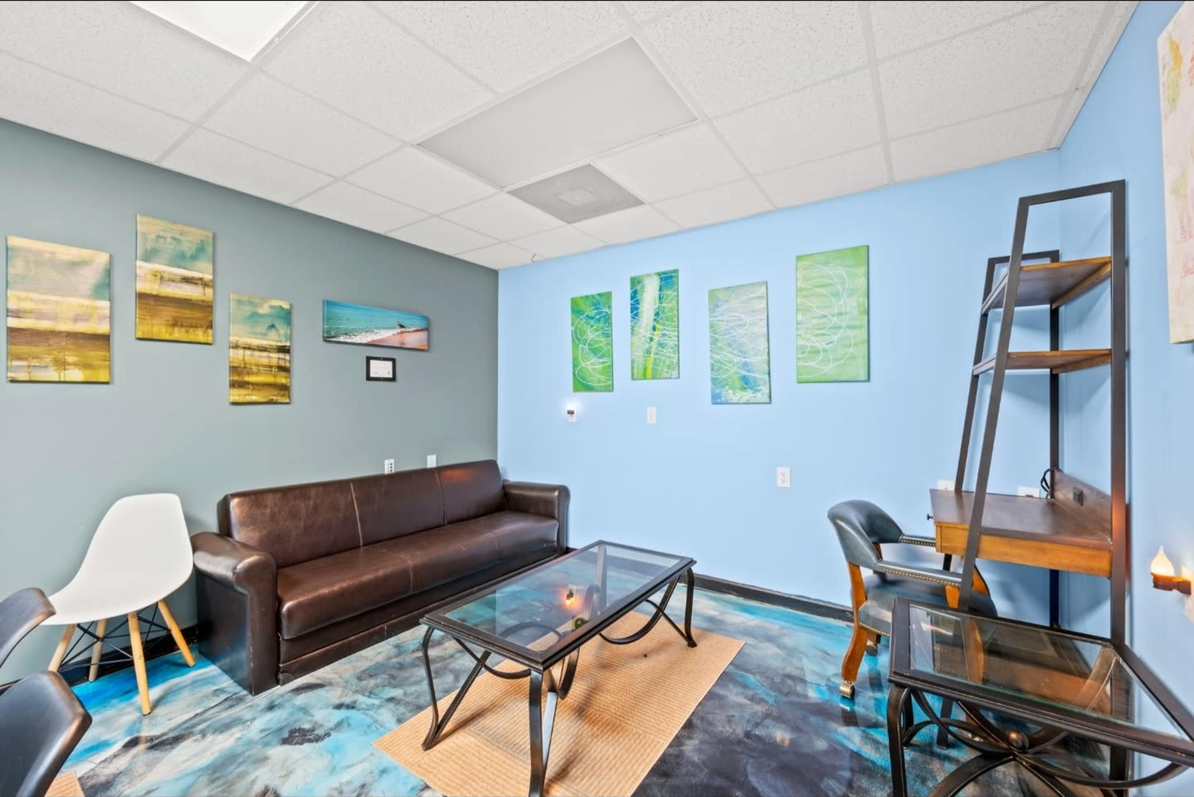 The image shows a cozy waiting area with a brown leather sofa, a glass coffee table, and several pieces of artwork on the walls in shades of blue and green.