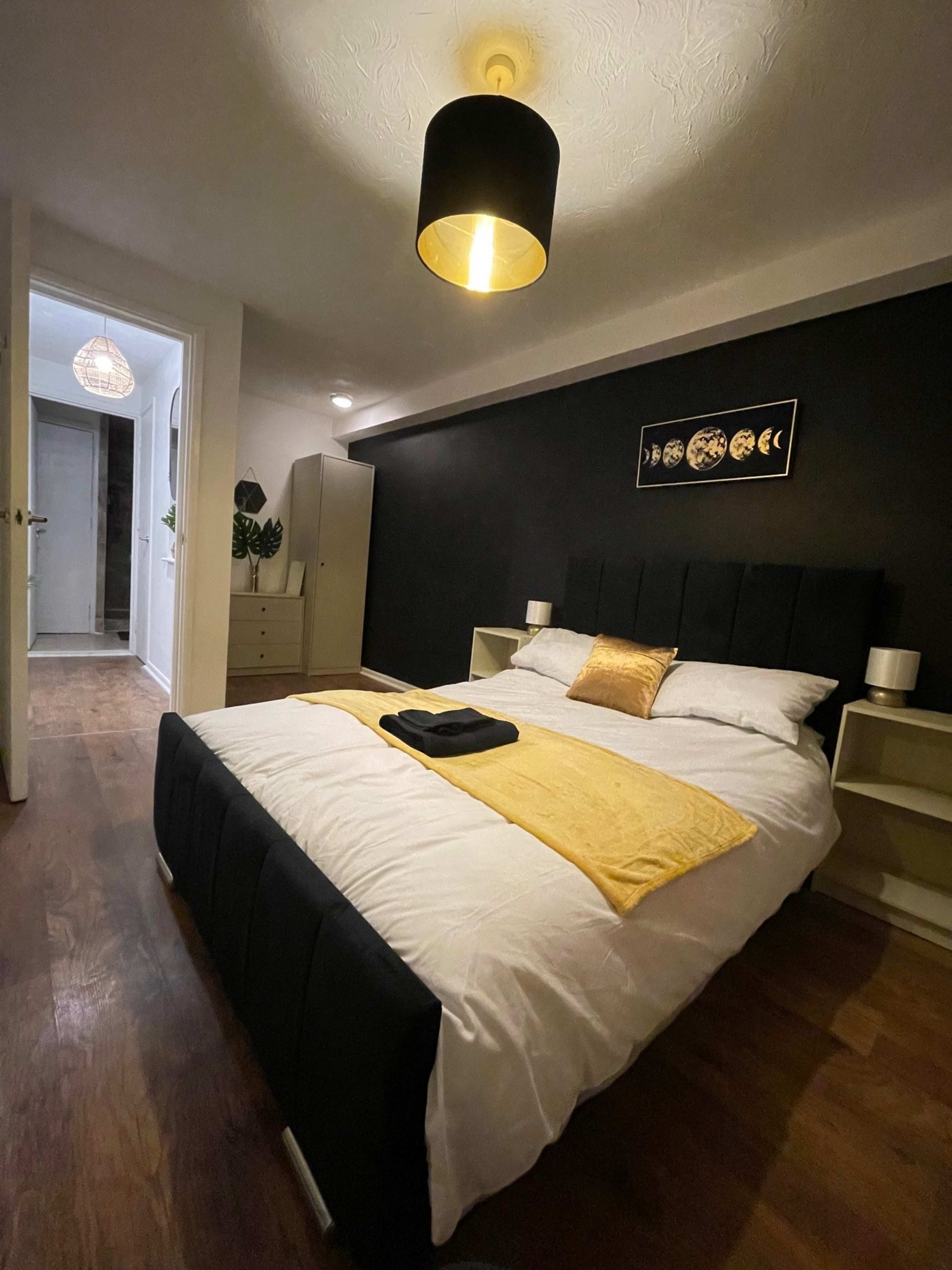 The image shows a modern bedroom with a dark accent wall, a double bed adorned with white linens and a yellow throw, along with a nightstand on each side and a wardrobe in the background.