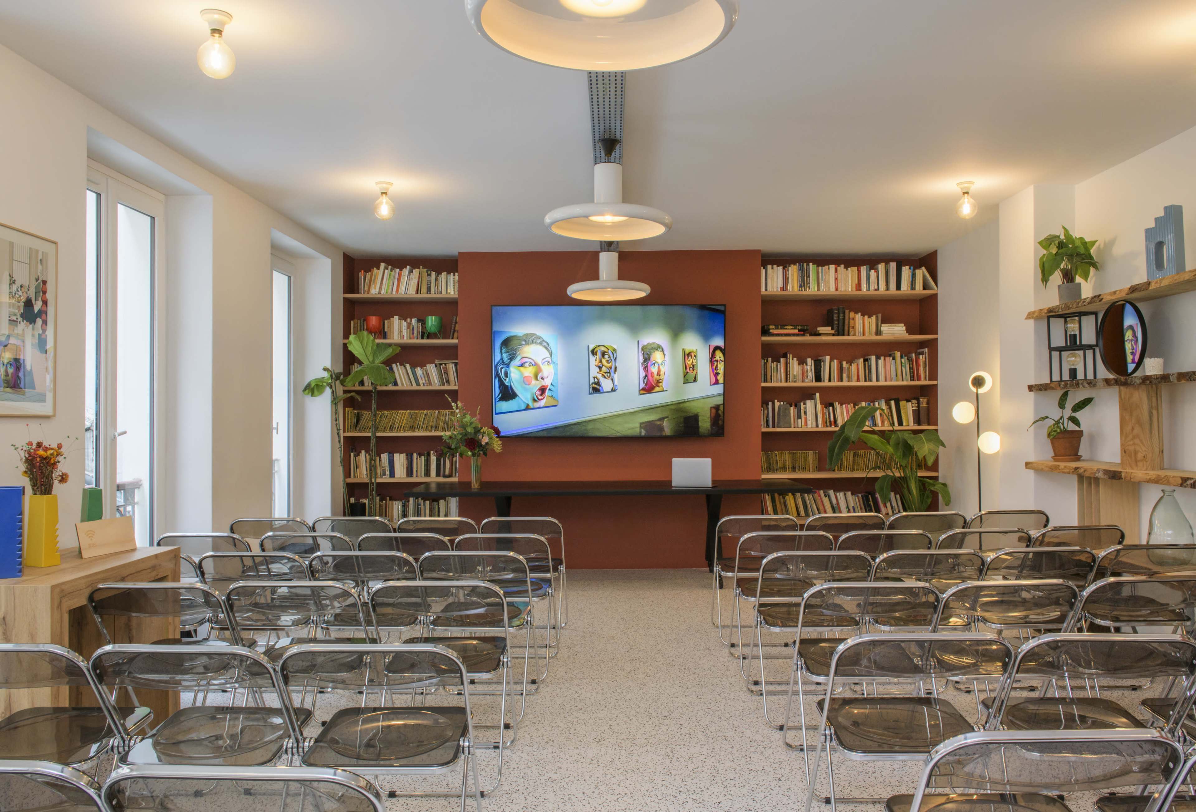 The image shows a modern meeting room with transparent chairs facing a screen displaying colorful artwork, surrounded by bookshelves and plants.