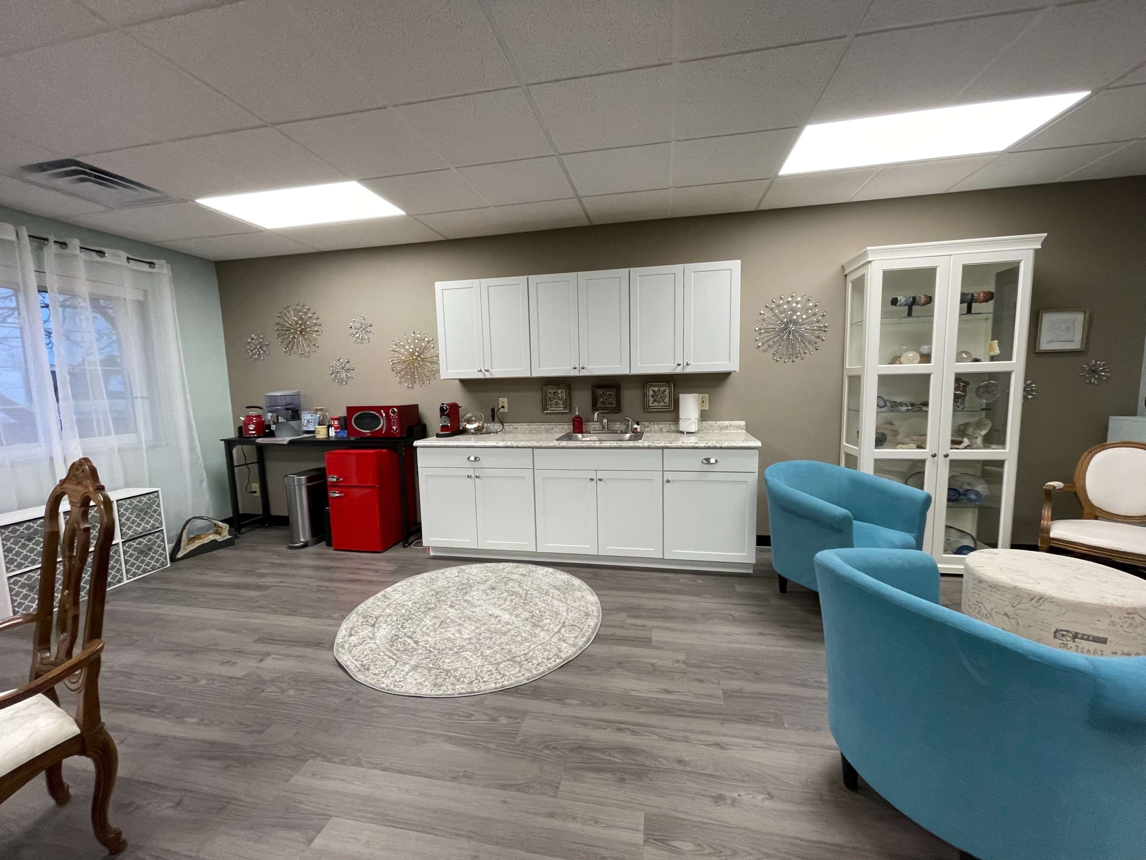A well-lit room featuring a kitchenette with white cabinets, a red mini refrigerator, and a display cabinet, along with light blue chairs and a round area rug.