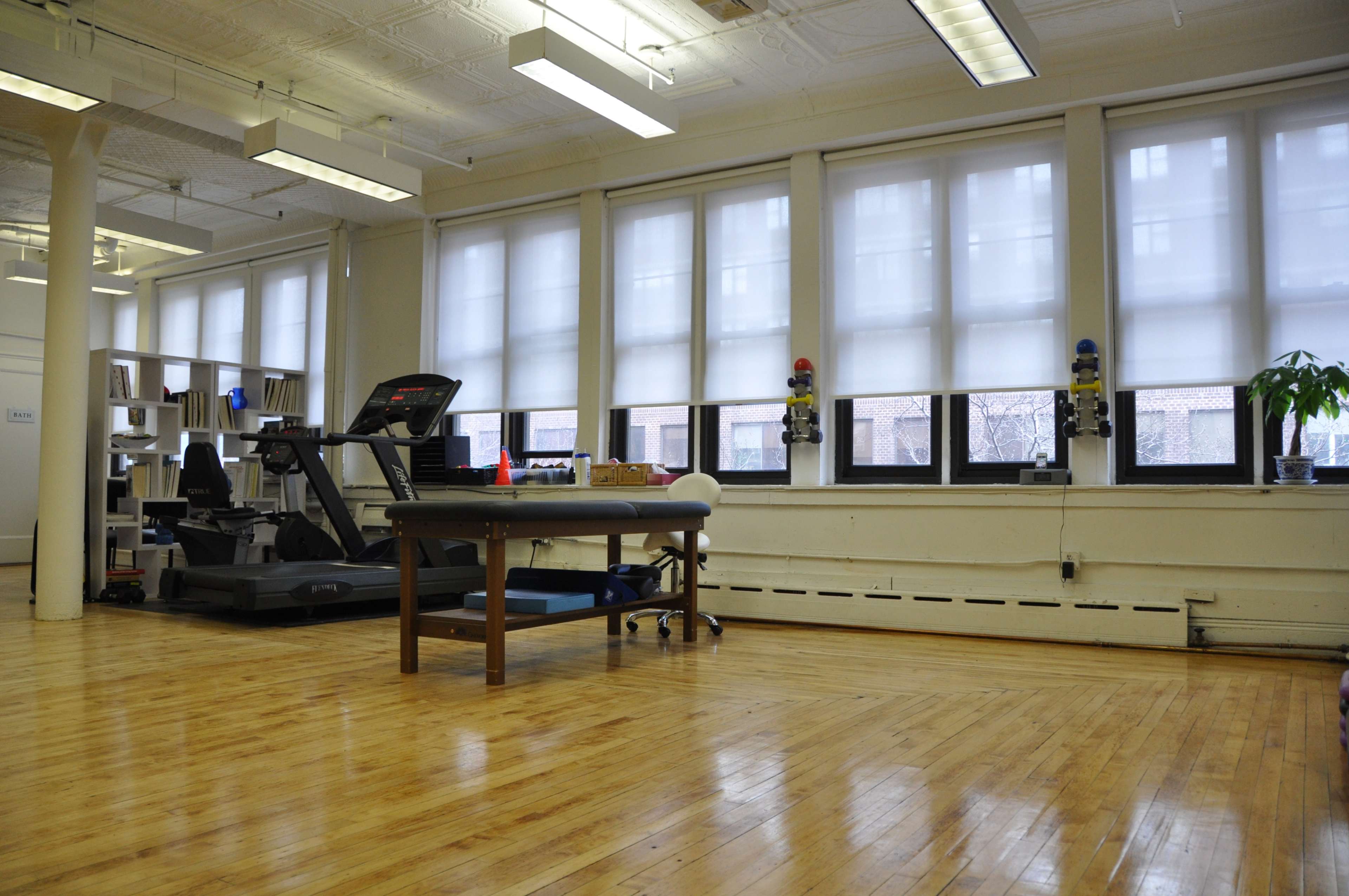 A spacious room with wooden floors, featuring a treadmill, a table, and large windows with blinds.