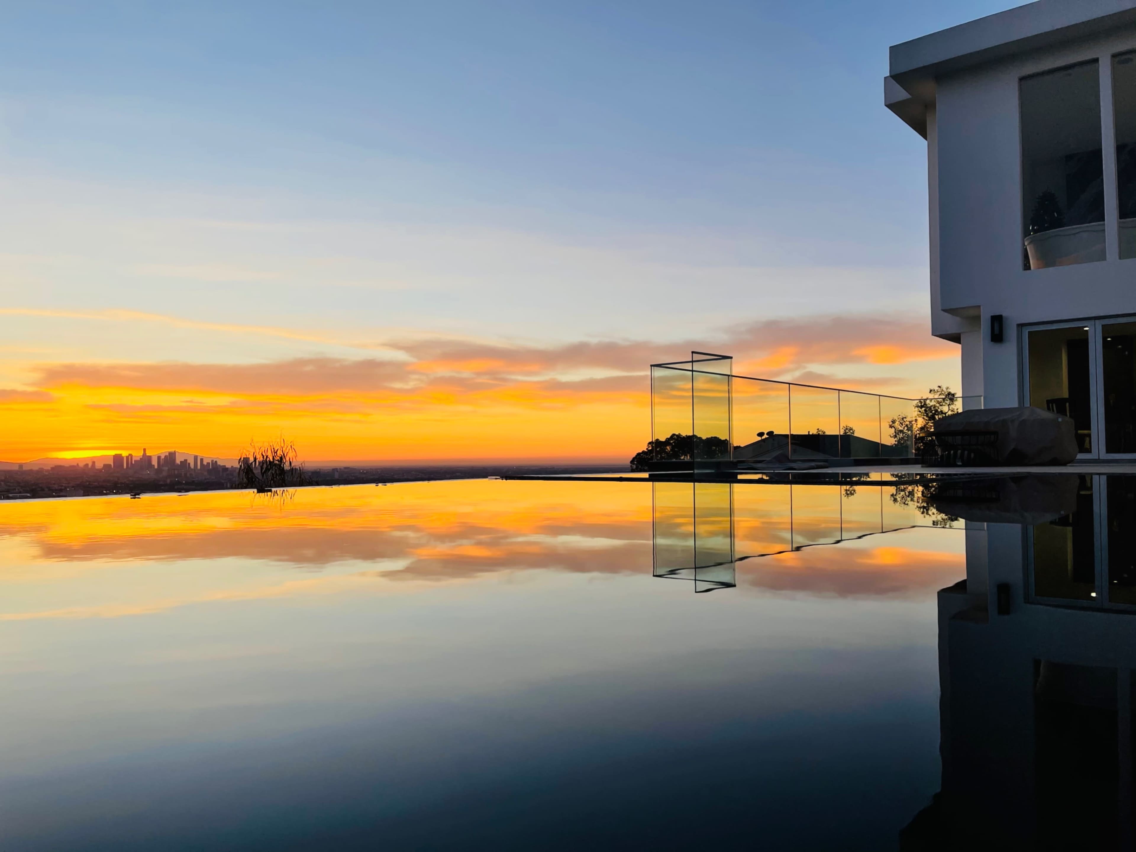 The image shows a luxurious modern home with a reflective infinity pool at sunset, casting vibrant colors across the sky and water.