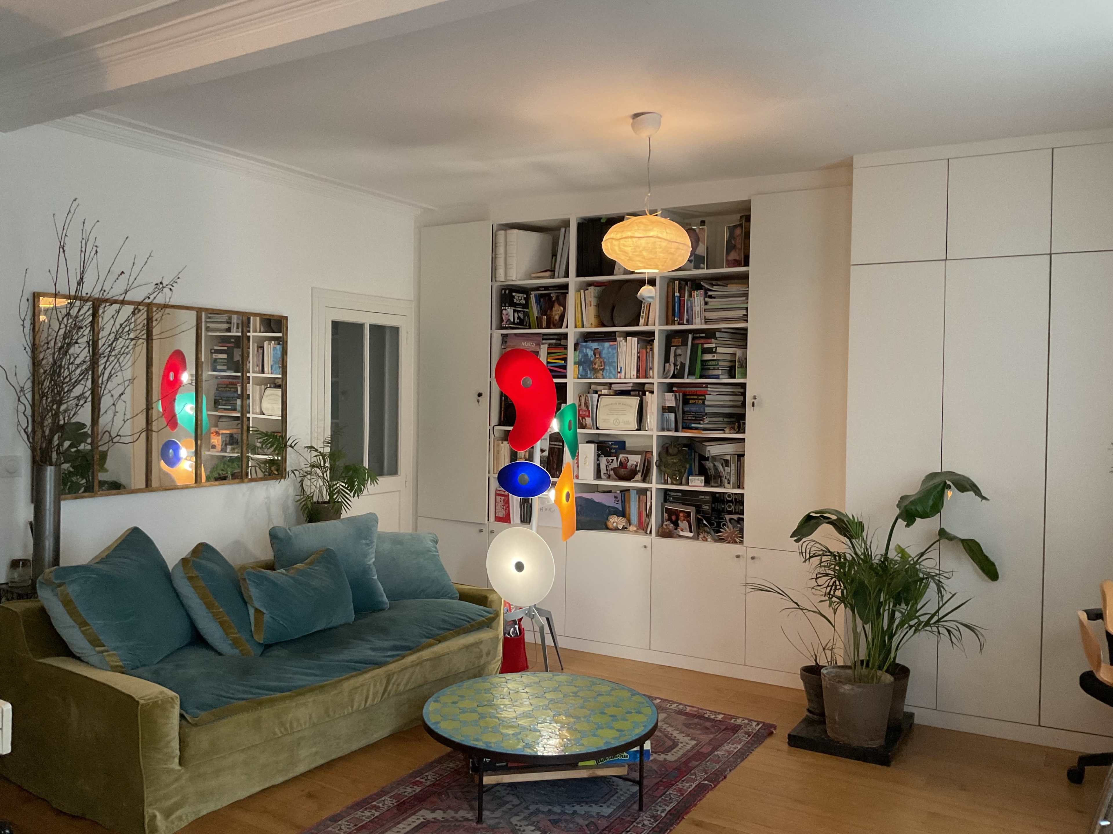 The image shows a cozy living room with a green velvet couch, a round coffee table, a plant, and shelves filled with various books and items.