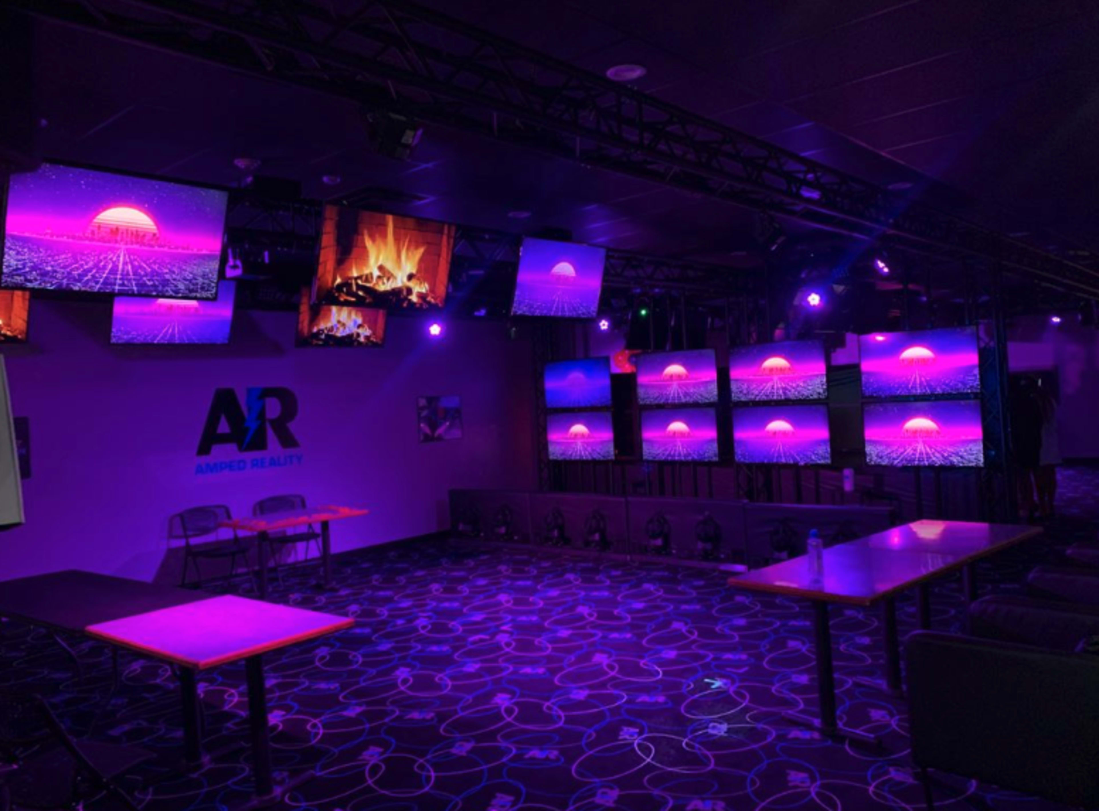 A dimly lit entertainment space featuring multiple screens that display vibrant sunset graphics, with tables and chairs arranged in the room.