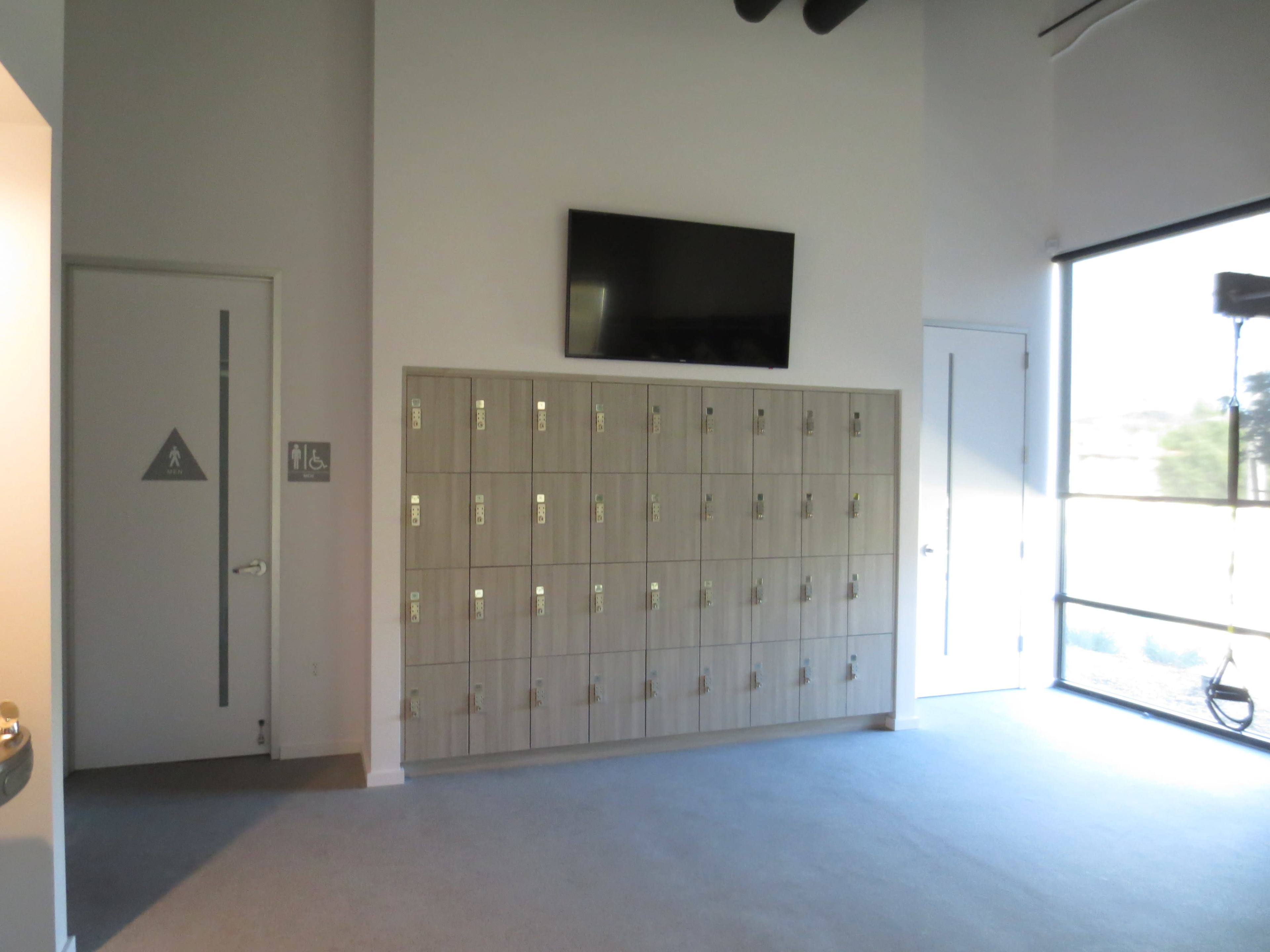 The image shows a room with a wall-mounted TV, a row of lockers, and a restroom sign on the door, illuminated by natural light from a large window.