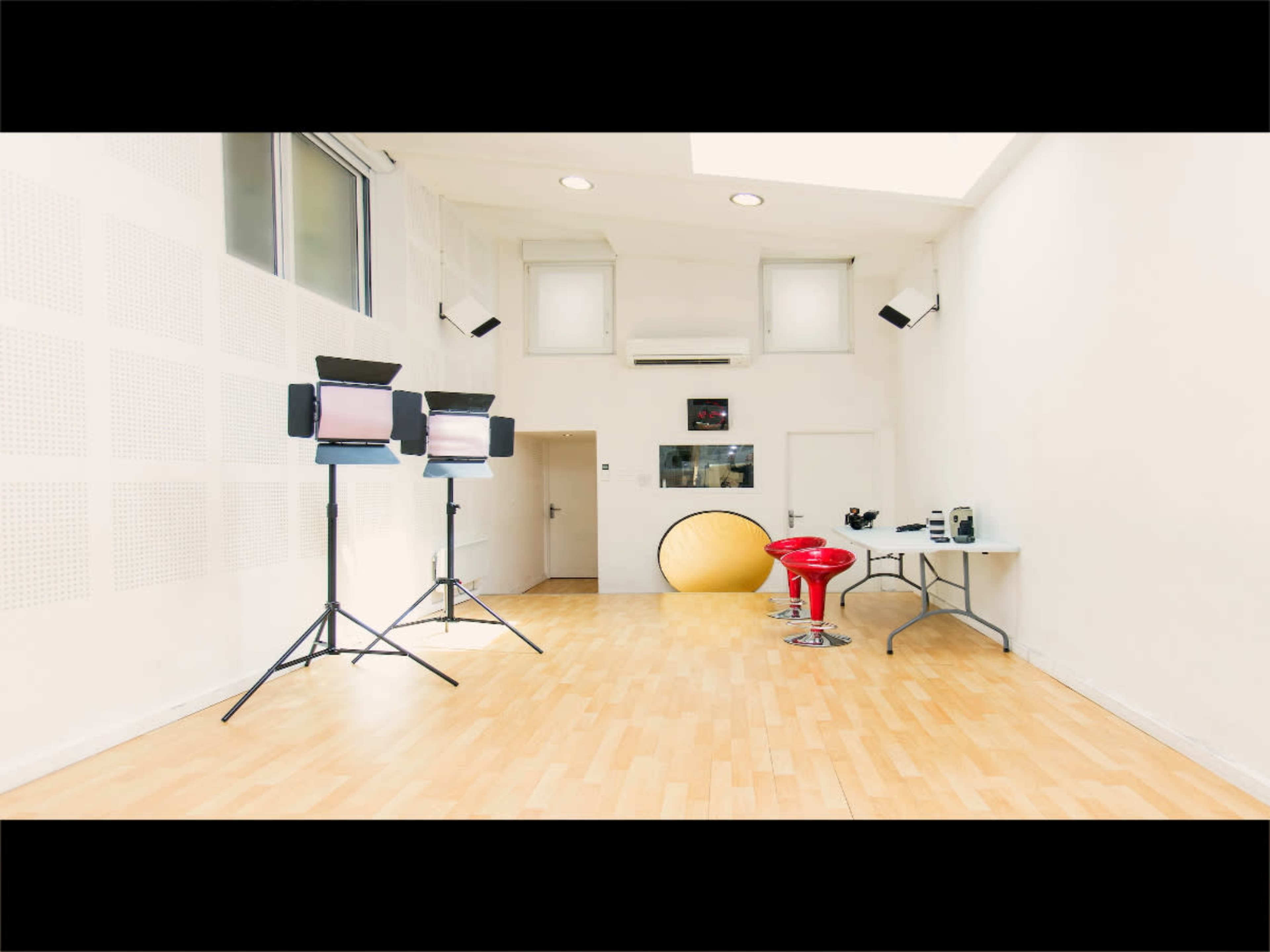 A brightly lit studio space featuring two lighting stands, a round yellow backdrop, a small table with equipment, and red bar stools.
