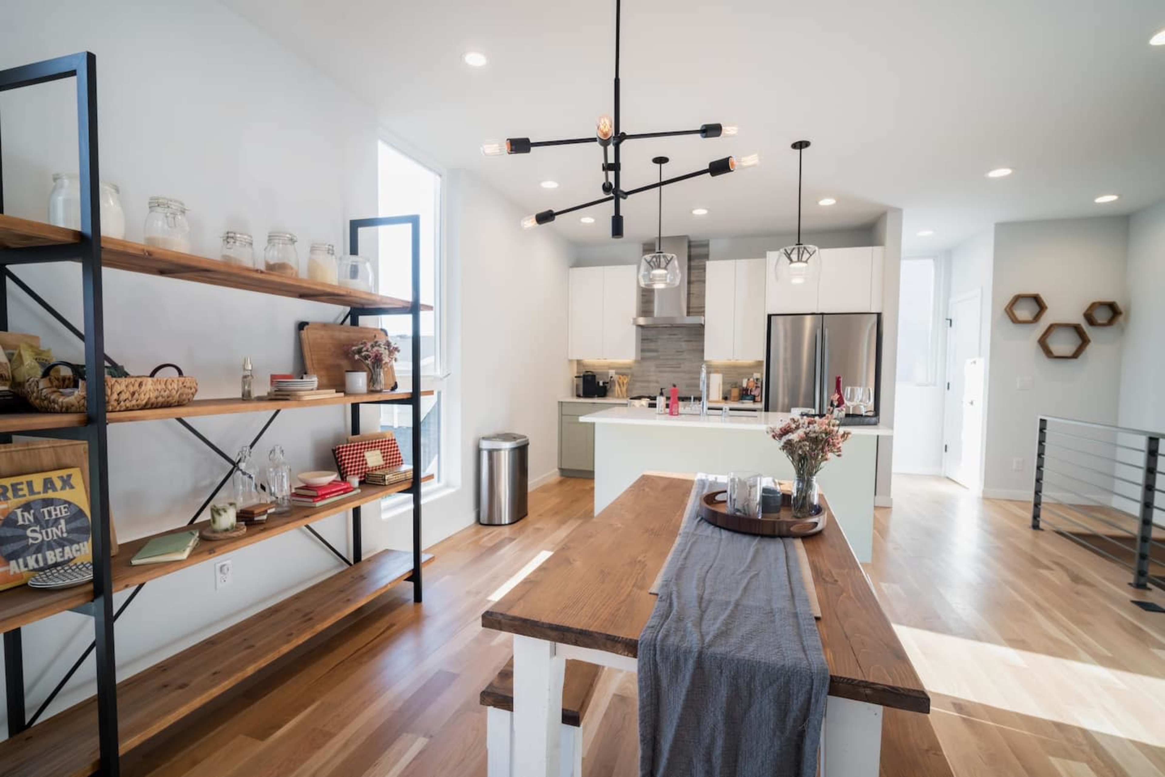 A modern kitchen with a wooden table, a black shelving unit, stainless steel appliances, and bright natural light from large windows.