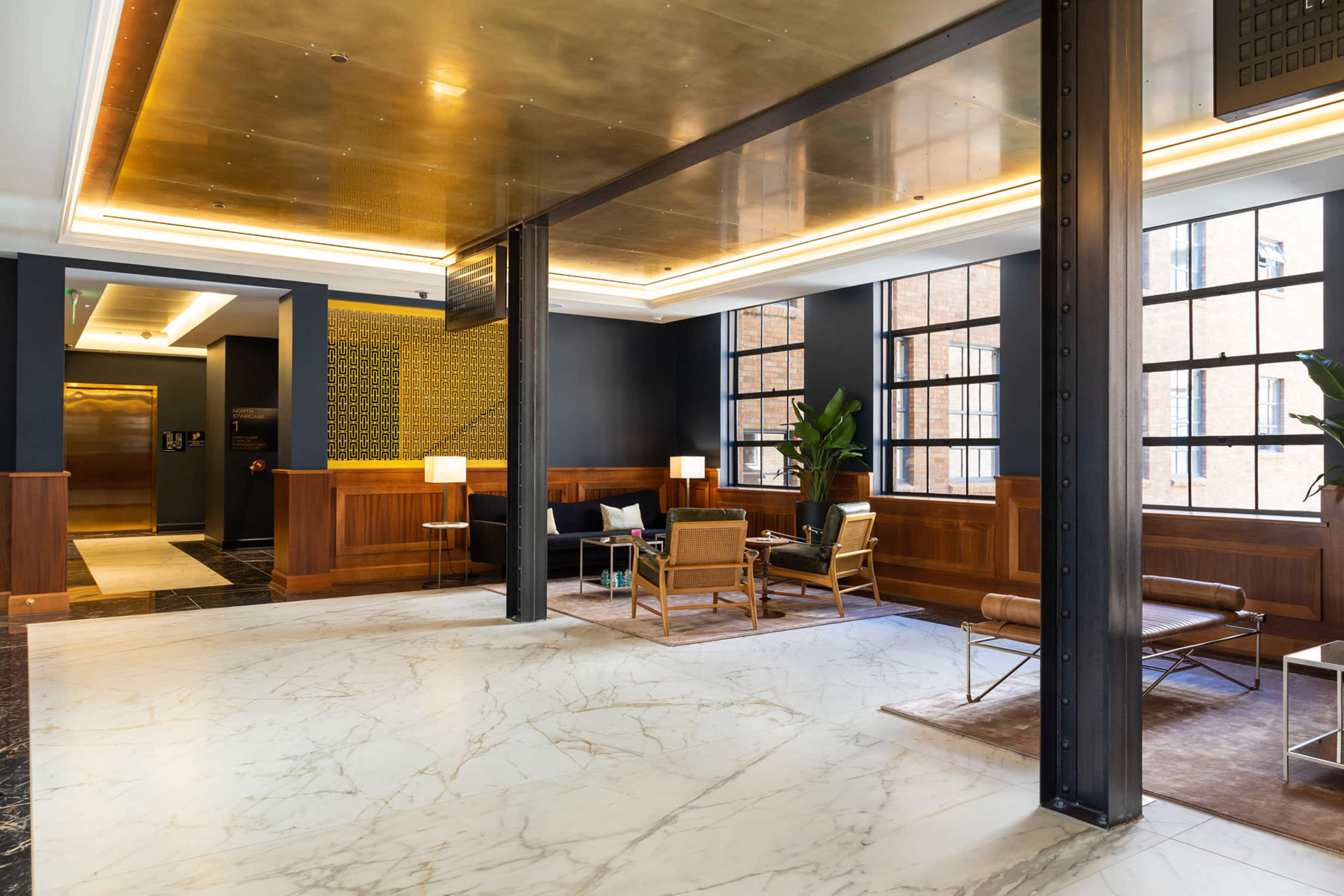 A spacious lobby with polished marble floors, wooden paneling, modern seating, and large windows allowing natural light to fill the area.