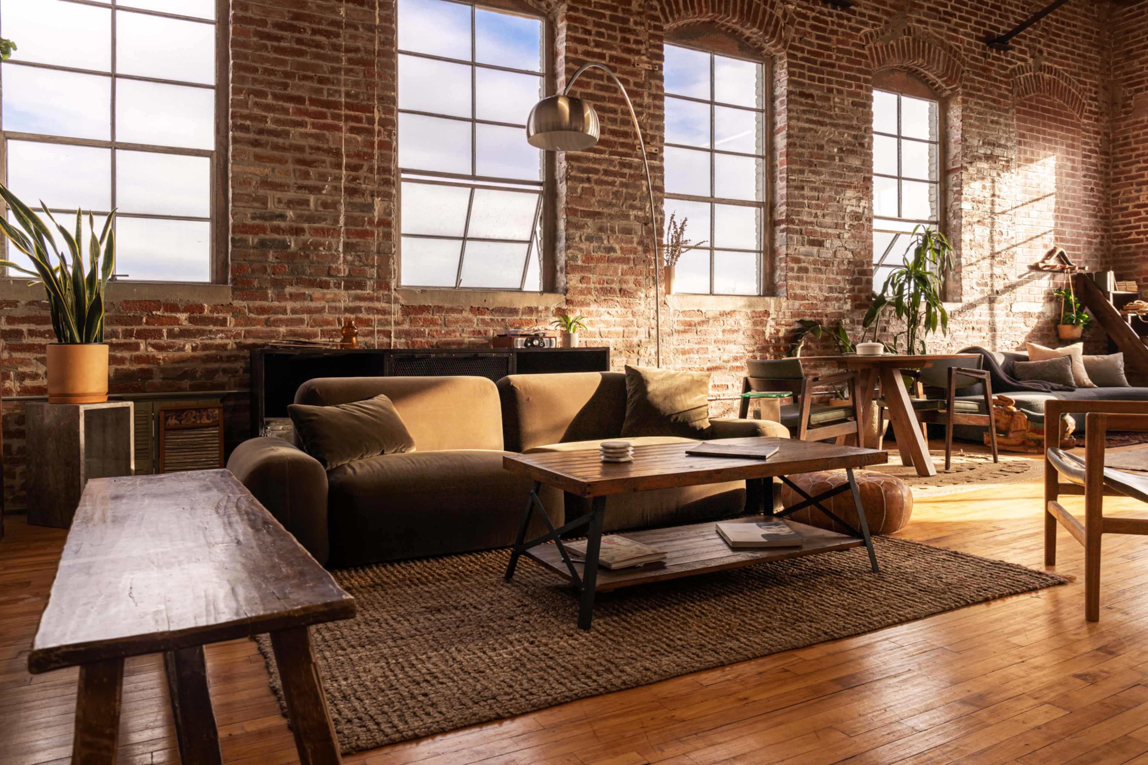 A spacious living room features exposed brick walls, large windows, and a mix of modern and rustic furniture.