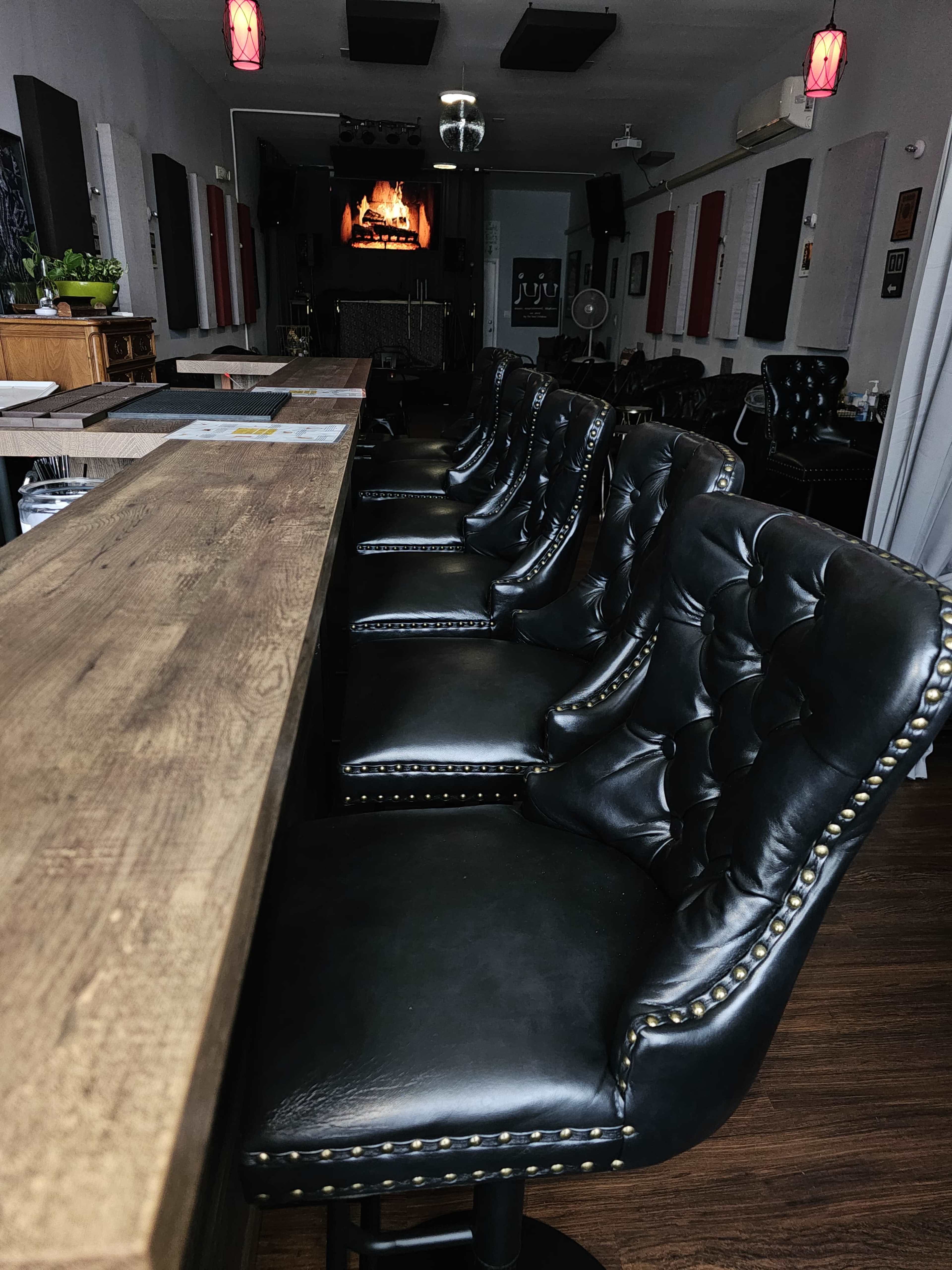 A row of black leather bar stools with tufted backs lines a wooden bar in a dimly lit establishment.