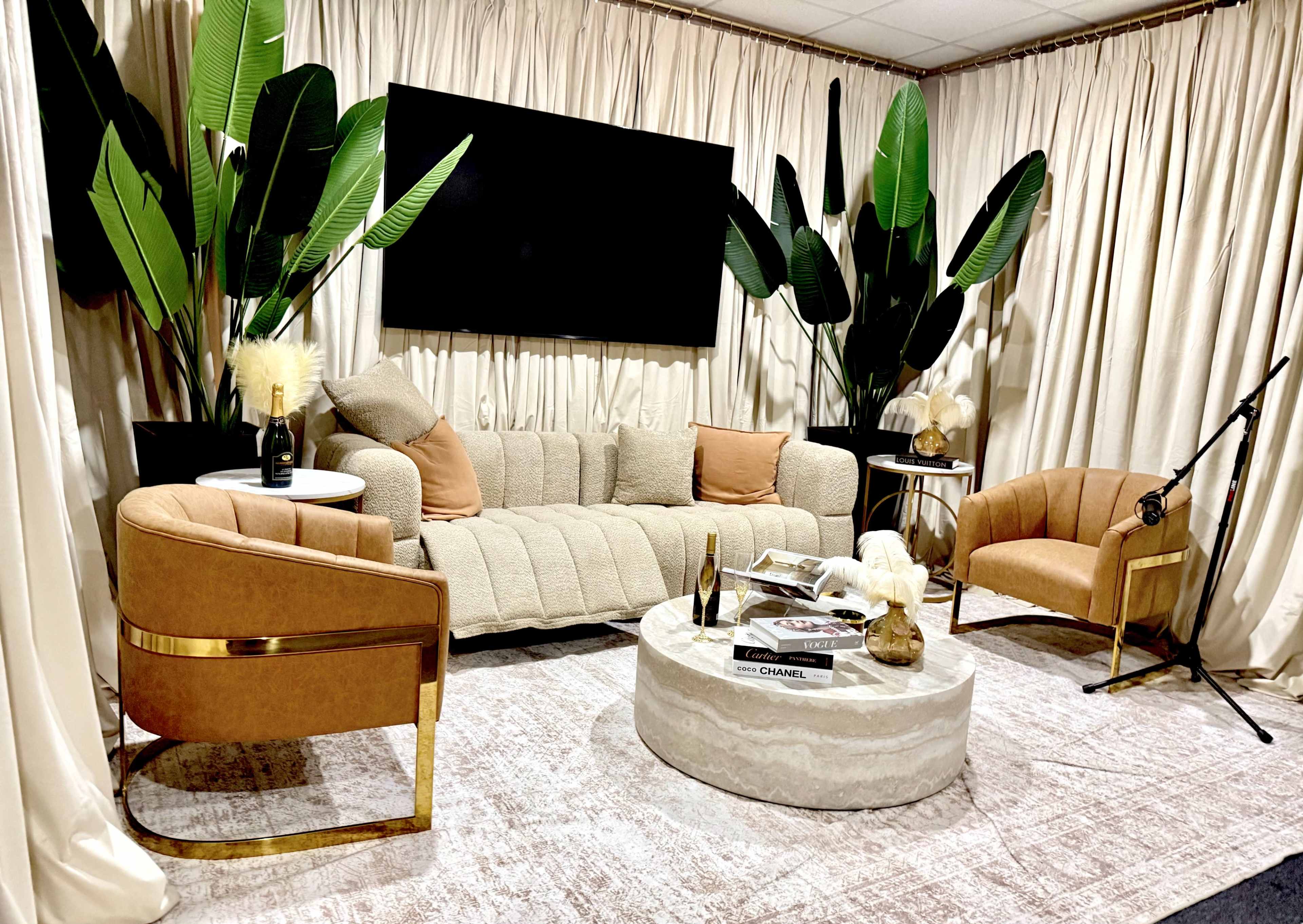 The image features a cozy living room setup with a tufted sofa, two upholstered chairs, a circular coffee table, and large leafy plants, all set against beige curtains and a mounted television.