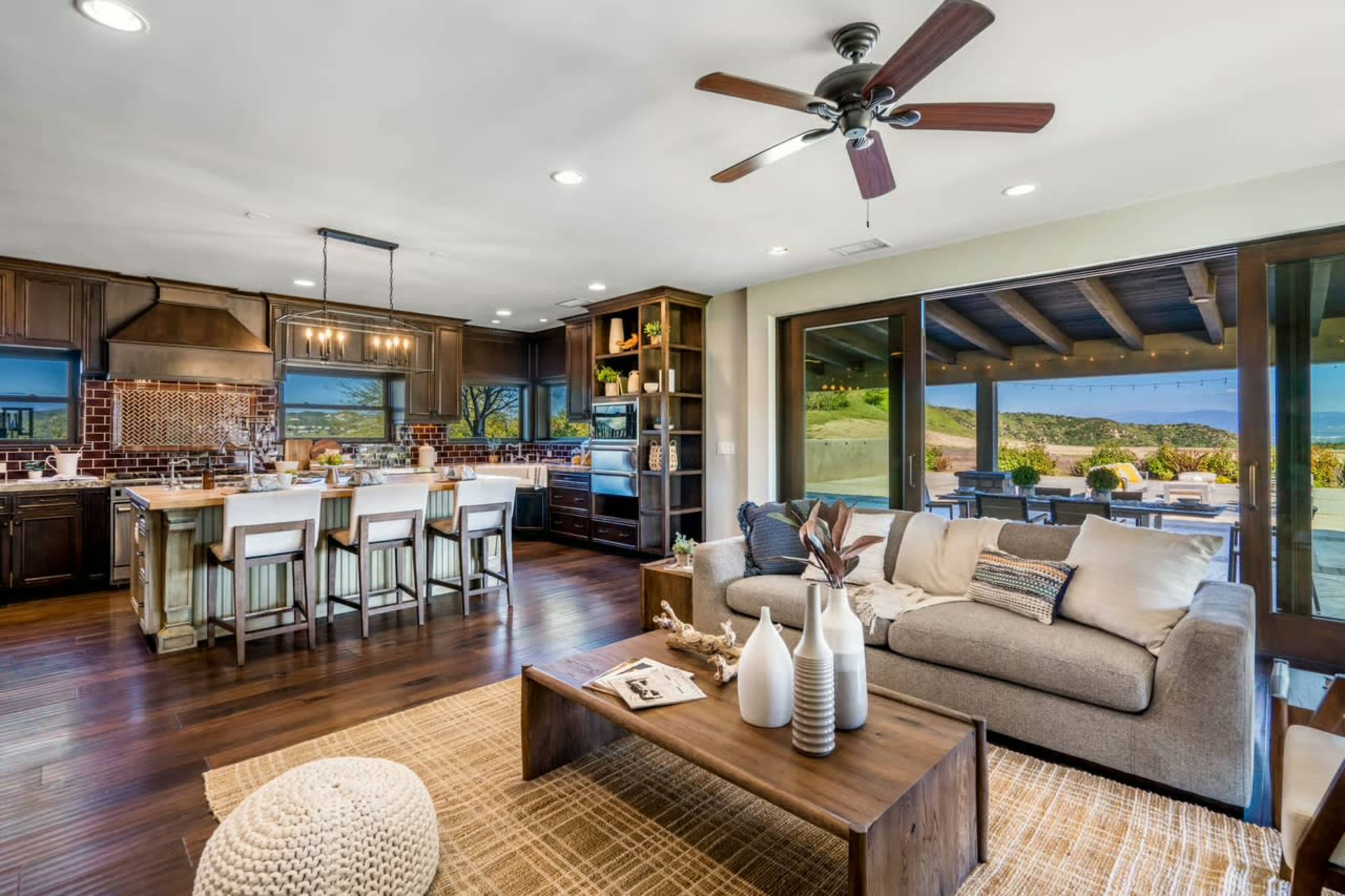 The image shows a modern kitchen and living area with wooden cabinets, a large island, and a comfortable seating arrangement, all overlooking a scenic outdoor patio.